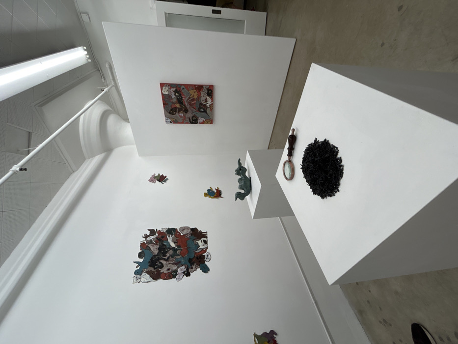 The scene is an interior view of an art gallery located in the city of Los Angeles, United States of America. The environment features white walls, a white ceiling with exposed fluorescent light fixtures, and a light gray concrete floor. A fluted column capital is visible near the ceiling on the right. A single human foot, wearing a dark shoe, is partially visible in the bottom left corner.
On the left wall, a large rectangular artwork composed of numerous overlapping, stylized figures in various colors (black, brown, red, blue, grey, white) is displayed. Below and to its right, several smaller, individual cut-out figures of similar style are affixed to the wall.
Two white square display pedestals are positioned centrally. The rear pedestal supports a greenish-blue sculpted object, approximately 18 inches long, resembling a reclining creature with stylized features. The foremost pedestal displays a conical pile of small, irregular black granular material, roughly 6 inches in diameter. Adjacent to this pile, a magnifying glass with a dark wooden handle and a bronze-toned rim is placed.
Further into the room on the right, a freestanding white partition displays a square-format painting. This artwork features a red background densely populated with stylized figures and shapes, similar in style to the wall-mounted pieces, incorporating black, white, and grey elements. A white door is partially visible in an open doorway on the far right background.