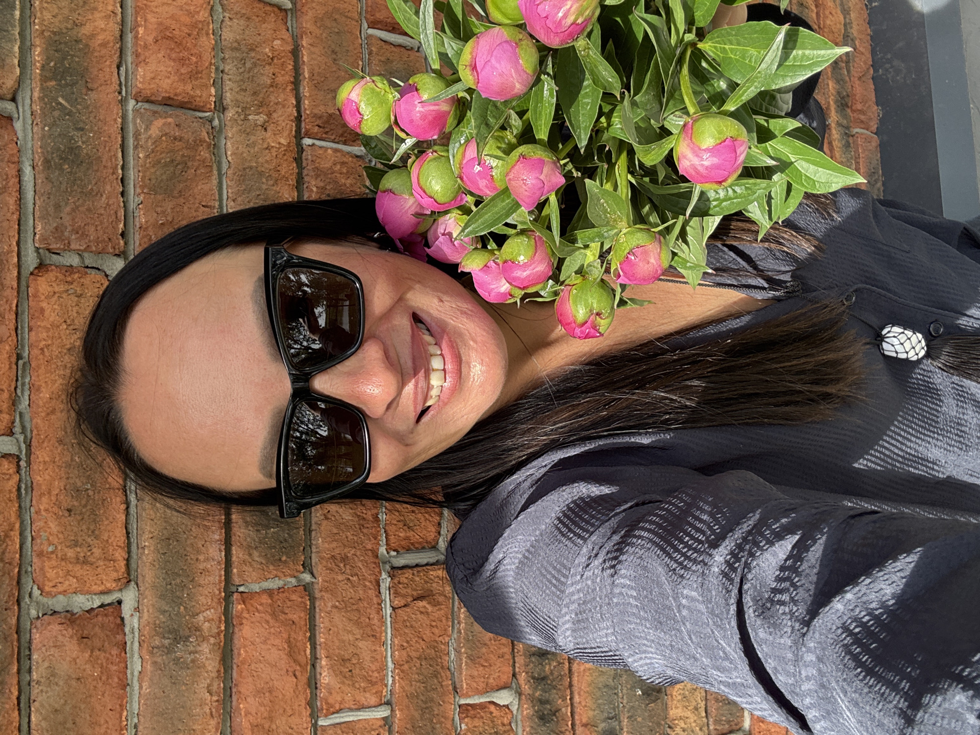 The image is a close-up selfie of a woman with dark hair wearing sunglasses and holding a bouquet of pink peonies. She is smiling and appears to be in good spirits. The background consists of a brick wall and a hint of a gray surface.
While the prompt mentioned Chaikovsky, Russia, there is no visible information in the image to confirm this location. The description remains focused on the observable details.