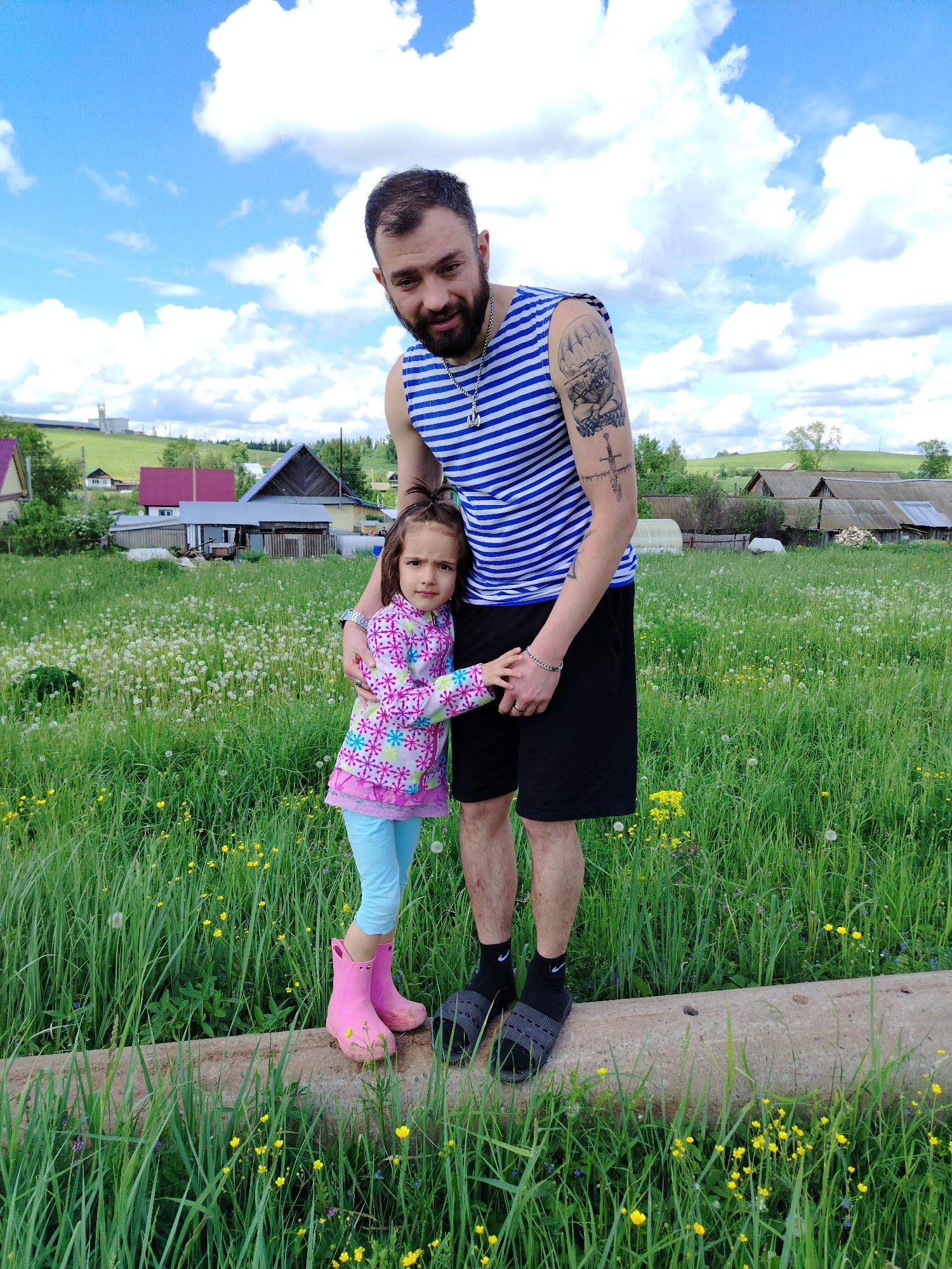 A full-shot photograph captures a man and a young girl in an outdoor setting in Гожан, Russia. The man stands behind the girl, his arm around her. He wears a blue and white striped tank top, black shorts, black socks with a white Nike logo, and gray sandals. A tattoo is visible on his left upper arm. He has dark hair and a beard. The girl, standing slightly in front of him, wears a long-sleeved, multicolored floral print shirt, light blue leggings, and pink rubber boots. They both stand on what appears to be a concrete beam.
The background features a field of tall green grass interspersed with dandelions and yellow wildflowers. Behind the field are several rural houses and outbuildings with varying roof colors and architectural styles. A green, rolling hill is visible in the distance, with a blue sky dotted with white clouds overhead. The overall lighting of the scene suggests it is daytime.