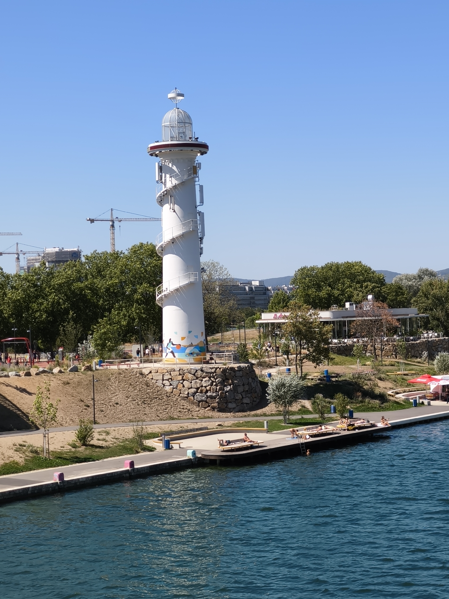 The image depicts a modern lighthouse with a spiral staircase and a glass-enclosed lantern room, situated on a small hill overlooking a body of blue water. The lighthouse is white and features a colorful mural near its base. In the foreground, a wooden pier extends into the water, where several individuals are sunbathing on loungers and one person is swimming. The pier is bordered by a walkway with colorful bollards and benches.
Behind the lighthouse, there is a park-like setting with trees, a paved path, and a stone wall. Further in the background, a modern building with a glass facade and an outdoor seating area, possibly a restaurant, is visible, surrounded by trees. Construction cranes and unfinished buildings are present on the horizon, suggesting ongoing development in the area. The sky is clear and blue.
This scene is located at the Donauplatte, a modern urban development area along the Danube River in Vienna, Austria, specifically near the U1 subway station "Donauinsel." This area is known for its recreational facilities and modern architecture.
