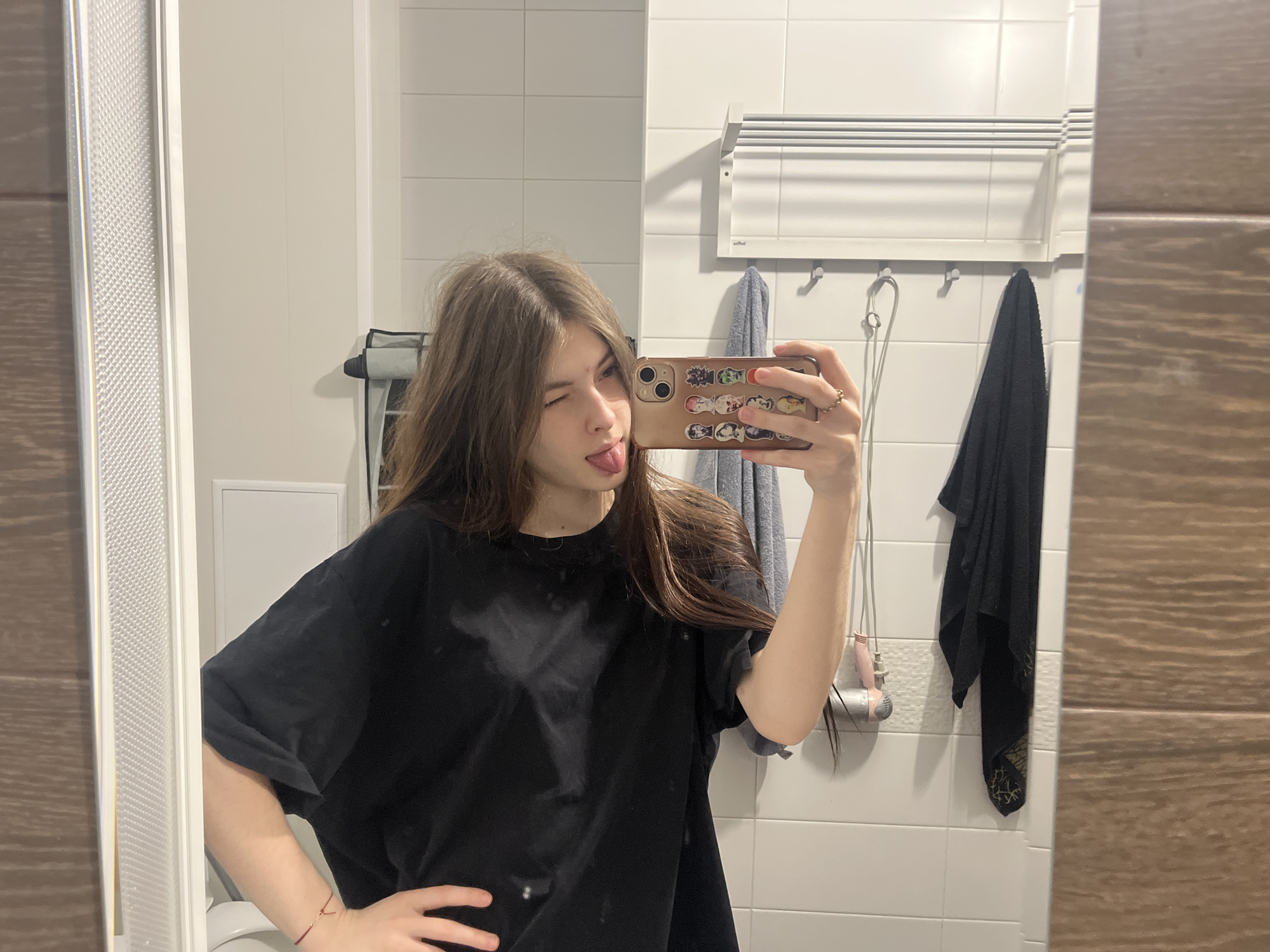A young woman is taking a selfie in a bathroom. She is wearing a black t-shirt and has long brown hair. She is sticking her tongue out and winking. She is holding a phone with stickers on the back. There is a towel rack with towels hanging on it in the background. There is also a hair dryer hanging on the wall. The location is likely a bathroom in an apartment in Saint Petersburg, Russia, based on the style of the bathroom tiles and fixtures.