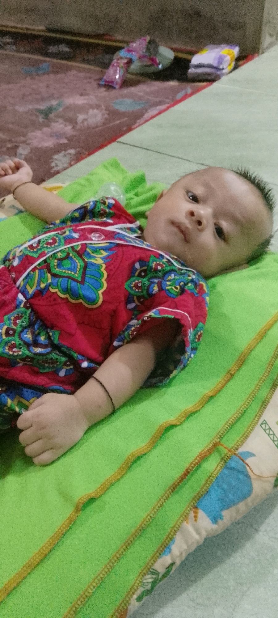 A baby is lying on its back on a bright green blanket. The baby is wearing a red and blue patterned garment. To the left of the baby, its left arm is extended and its hand is visible. A thin black bracelet is around the baby's left wrist. The background shows a patterned rug and what appears to be a wall and some packaged items on the rug. The scene appears to be indoors, likely in a residential setting in Lenggadai Hilir, Indonesia.