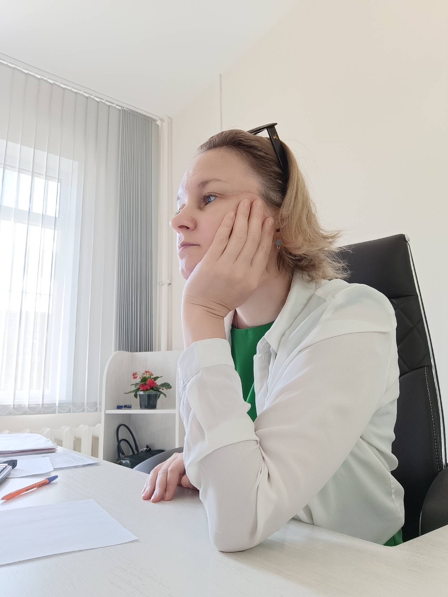 A middle-aged Caucasian woman with blonde hair pulled back and glasses perched on her head sits at a desk in an office. She is wearing a green shirt and a white button-down shirt. Her left hand is resting on her cheek as she looks off to the side, appearing pensive or bored. The desk is cluttered with papers, a calculator, and an orange pen. In the background, a window with vertical blinds lets in natural light. A small potted plant with red flowers sits on a shelf behind the desk, along with a black handbag. The overall environment suggests a typical office setting. This scene is located in Markovsky, Russia.