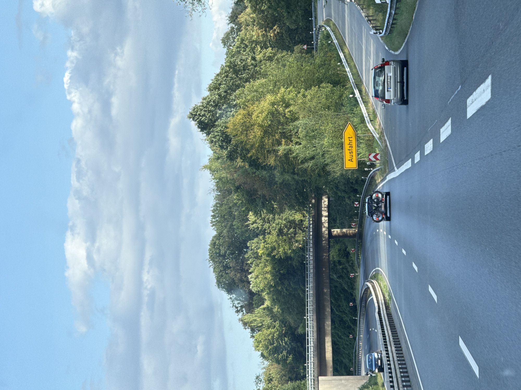 The image depicts a highway scene in Herzberg, Germany. The road is paved and has multiple lanes, marked with white dashed lines. On the right side of the road, a silver car with license plate "HH-MUA 223" is driving away from the camera. Further down the road, a yellow sign with the word "Ausfahrt" (German for "Exit") points to the right.
To the left of the highway, a concrete bridge spans across. A grey car is visible driving on the highway under the bridge. Beyond the highway, a dense forest with various shades of green trees lines the landscape. The sky above is blue with scattered white clouds. The overall scene suggests a travel context on a highway, with signs indicating an upcoming exit.