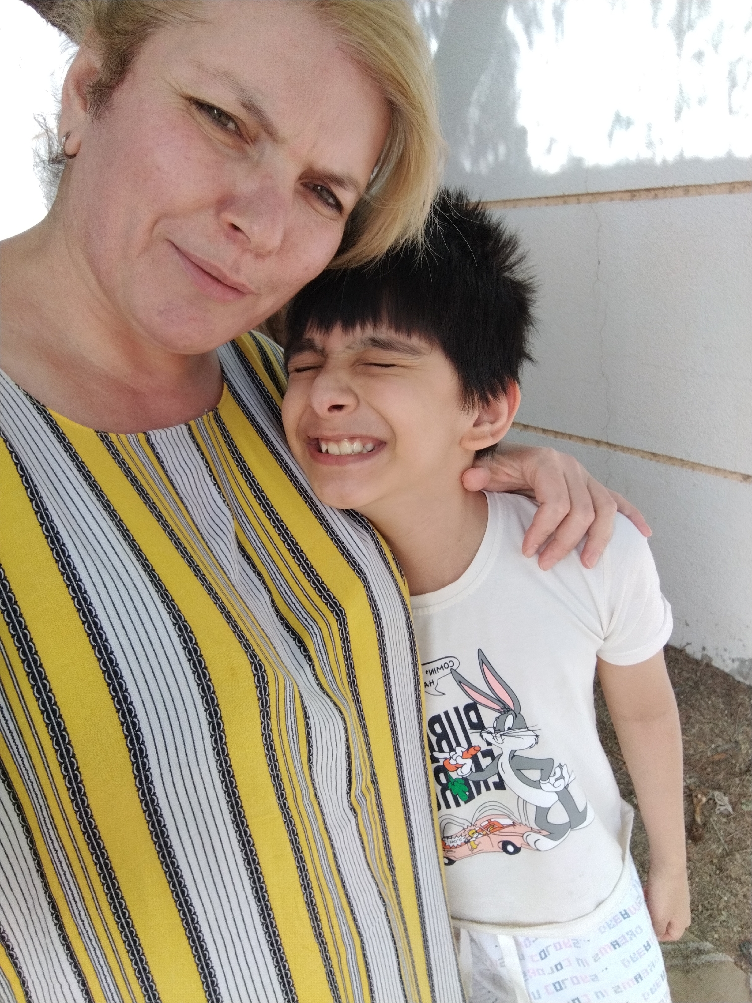 In Al Mas'oudi, United Arab Emirates, an indoor scene presents a woman and a child. The woman has short, blonde hair and is wearing a yellow and white striped top. The child, with dark hair, is embracing the woman with closed eyes and a smile. The child is wearing a white t-shirt with a Bugs Bunny cartoon graphic. The setting appears to be against a wall.