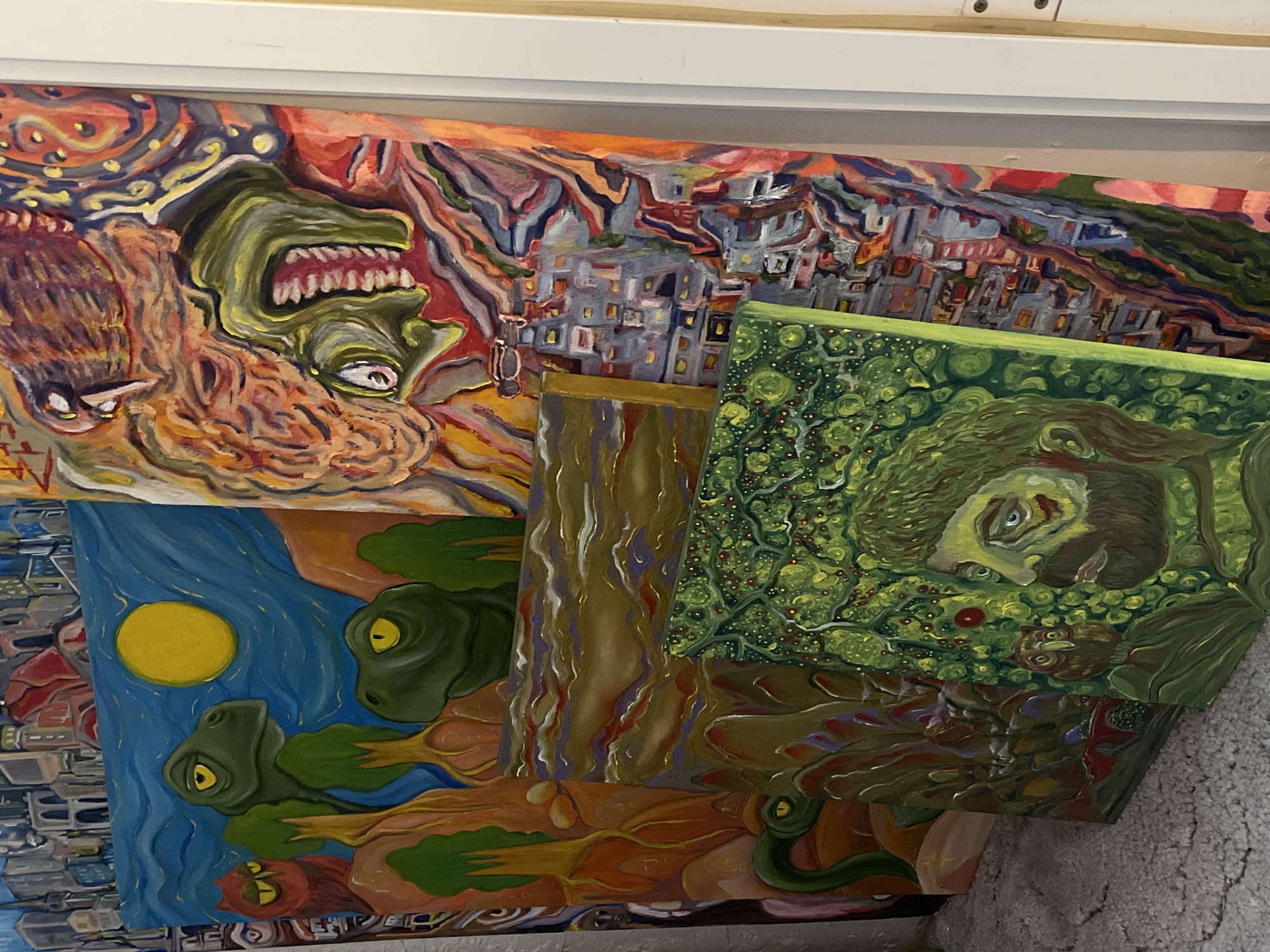 The image displays a collection of vibrant, surrealist paintings leaning against a wall, possibly in an artist's studio or exhibition space. The paintings feature a variety of subjects rendered in bold colors and expressive brushstrokes. One prominent painting depicts two dinosaur-like heads with yellow eyes, set against a blue sky with a large yellow sun. To the right of this, another large canvas shows a chaotic, abstract scene with distorted faces and a cityscape. In the foreground, a smaller painting features a man's profile surrounded by green foliage and small red fruits, with an owl visible near his shoulder. The floor appears to be carpeted with a textured, grey material. The paintings are layered and angled, suggesting they are being stored or displayed. There is no indication of the specific location in Krasnoyarsk, Russia.