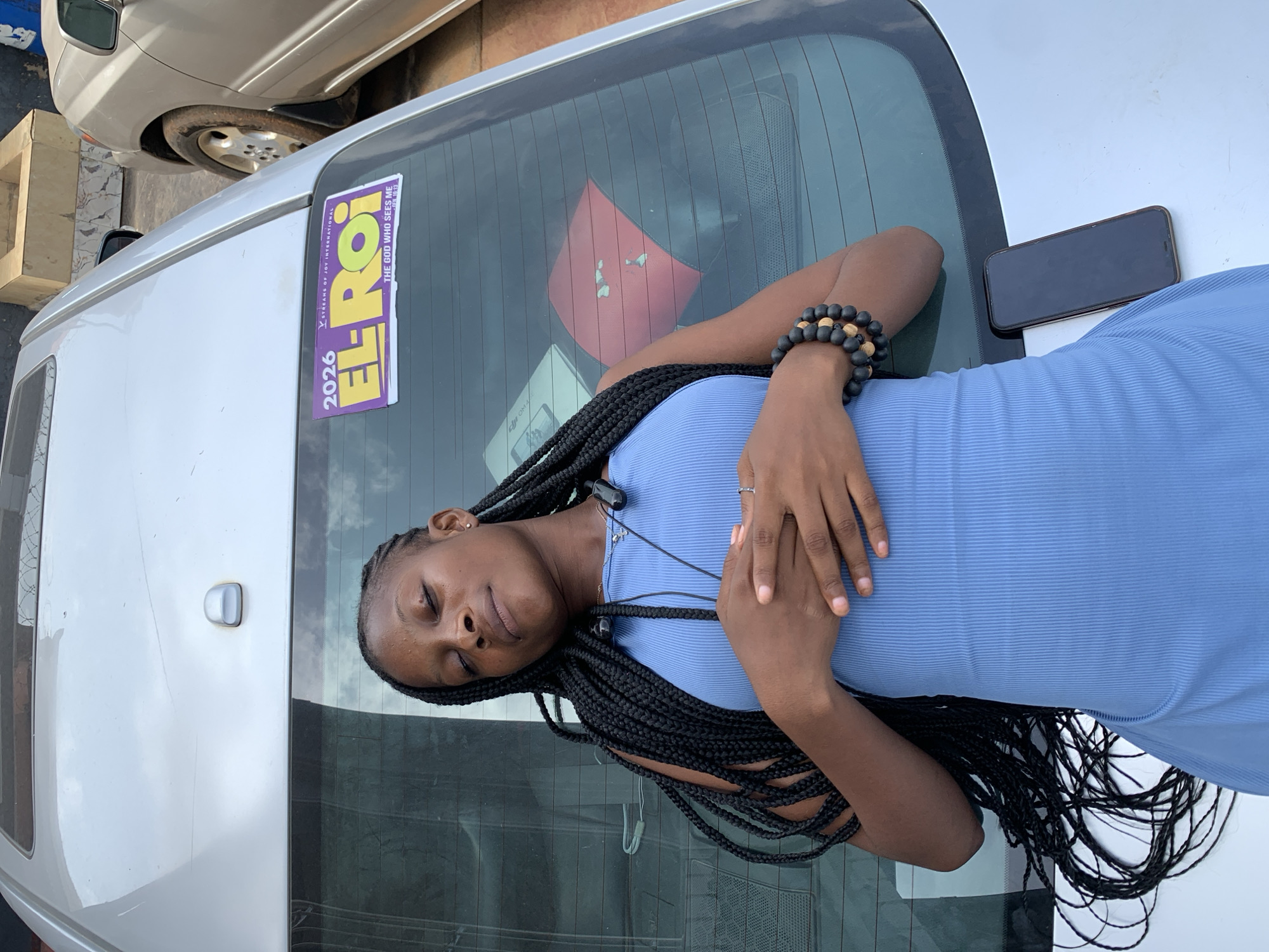 A young Black woman with long braids is lying on the hood of a silver car. She is wearing a blue dress and a beaded bracelet. A cell phone is next to her on the hood of the car. A sticker with the words "EL-ROI THE GOD WHO SEES ME" and "2026" on it is affixed to the car's rear window. In the background, there is another car and what looks like a building. The location is indicated by the "EL ROI" sticker to be the city of Ikorodu, Nigeria.