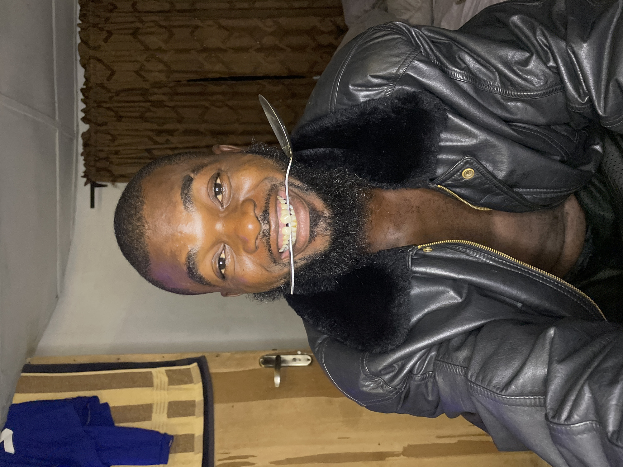 This image features an adult Black male subject, centrally positioned and looking directly at the viewer, within an indoor domestic setting located in Jos, Nigeria. The man has short dark hair, a full dark beard, and a mustache. He is smiling widely, revealing his teeth, and holds a silver-colored spoon horizontally between his upper and lower front teeth. He is wearing an unzipped black faux leather jacket with a dark faux fur collar, exposing his bare chest.
To the subject's left, a light brown wooden door with a metallic handle and lock is visible. Above and to the left of the door, a blue long-sleeved garment with a white shoulder accent is draped, next to a vertically striped fabric item featuring alternating light brown and dark brown bands. To the subject's right, dark brown, textured curtains are drawn, and a dark curtain rod is visible above them. The ceiling is white with visible panel seams. The overall illumination is warm and appears to be from an artificial light source.