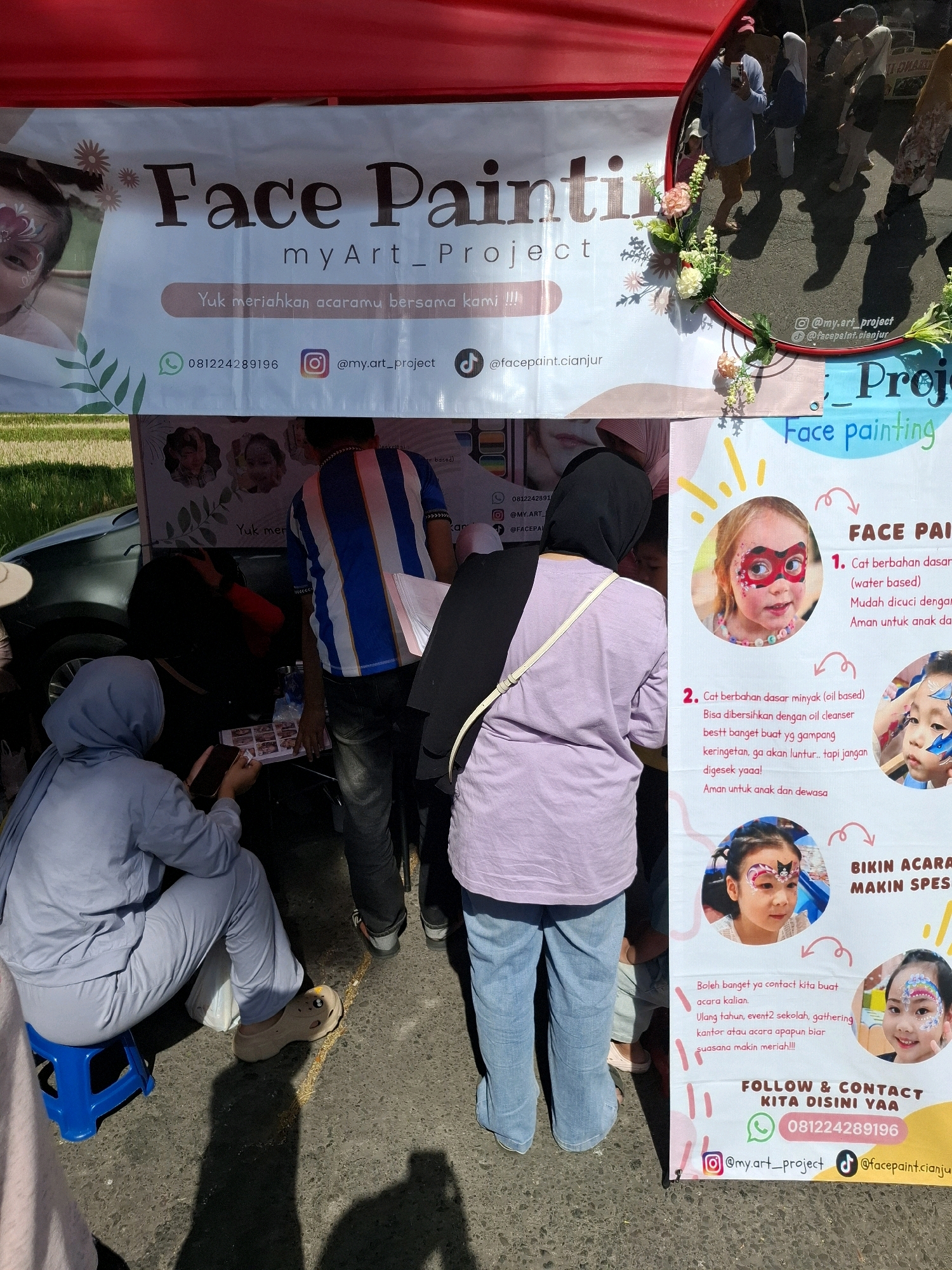 This is a medium shot, eye-level, outdoor, daytime photograph taken in Karangtengah, Indonesia. The image depicts a face painting stall at what appears to be a local event or market. A white banner with red and brown text is prominently displayed, advertising "Face Painting" services by "myArt_Project". Below this, a caption reads "Yuk meriahkan acaramu bersama kami !!!" (Let's liven up your event with us!!!). Contact information including a phone number (081224289196), Instagram handle (@my.art_project), and TikTok handle (@facepaint.cianjur) are visible on the banner.
In the foreground, a person wearing a light blue hijab and matching pants is seated on a blue stool, holding a mobile phone. To their right, another person in a light purple top and blue jeans stands, facing away from the camera. Behind them, other individuals are partially visible. A dark colored car is parked to the left of the scene.
To the right of the main banner, a second, colorful banner with yellow and pink elements is visible. This banner features images of children with face paint and provides information about the types of face paint used: water-based and oil-based, along with descriptions of their properties and safety. A call to action is present, encouraging people to contact them for events like birthdays, school events, gatherings, or office functions. The bottom of this banner reiterates the contact information.
The ground appears to be asphalt or a similar paved surface, with shadows cast by people and objects. The overall atmosphere suggests a lively community event.