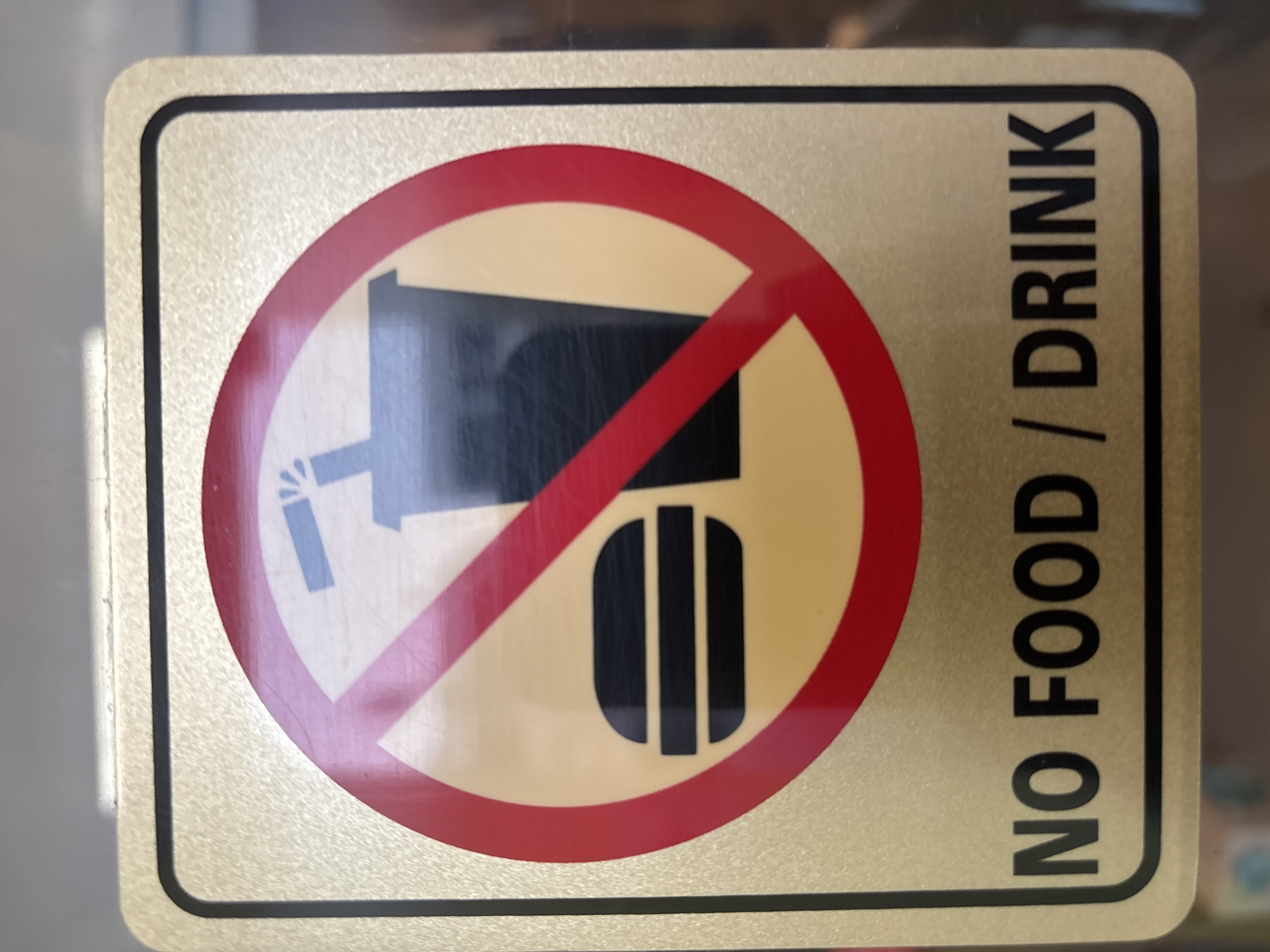 A close-up, eye-level shot shows a rectangular "NO FOOD / DRINK" sign with a metallic gold background and a black border. In the center of the sign, a red circle with a diagonal red line through it indicates prohibition. Inside the circle, a stylized black hamburger is on the left, and a black cup with a straw and a splash graphic is on the right. The text "NO FOOD / DRINK" is printed in bold black letters below the prohibition symbol. The sign appears to be affixed to a glass surface, with a faint reflection visible. The background behind the sign is blurred and appears to be an indoor environment. The location is identified as Pekanbaru, Indonesia.