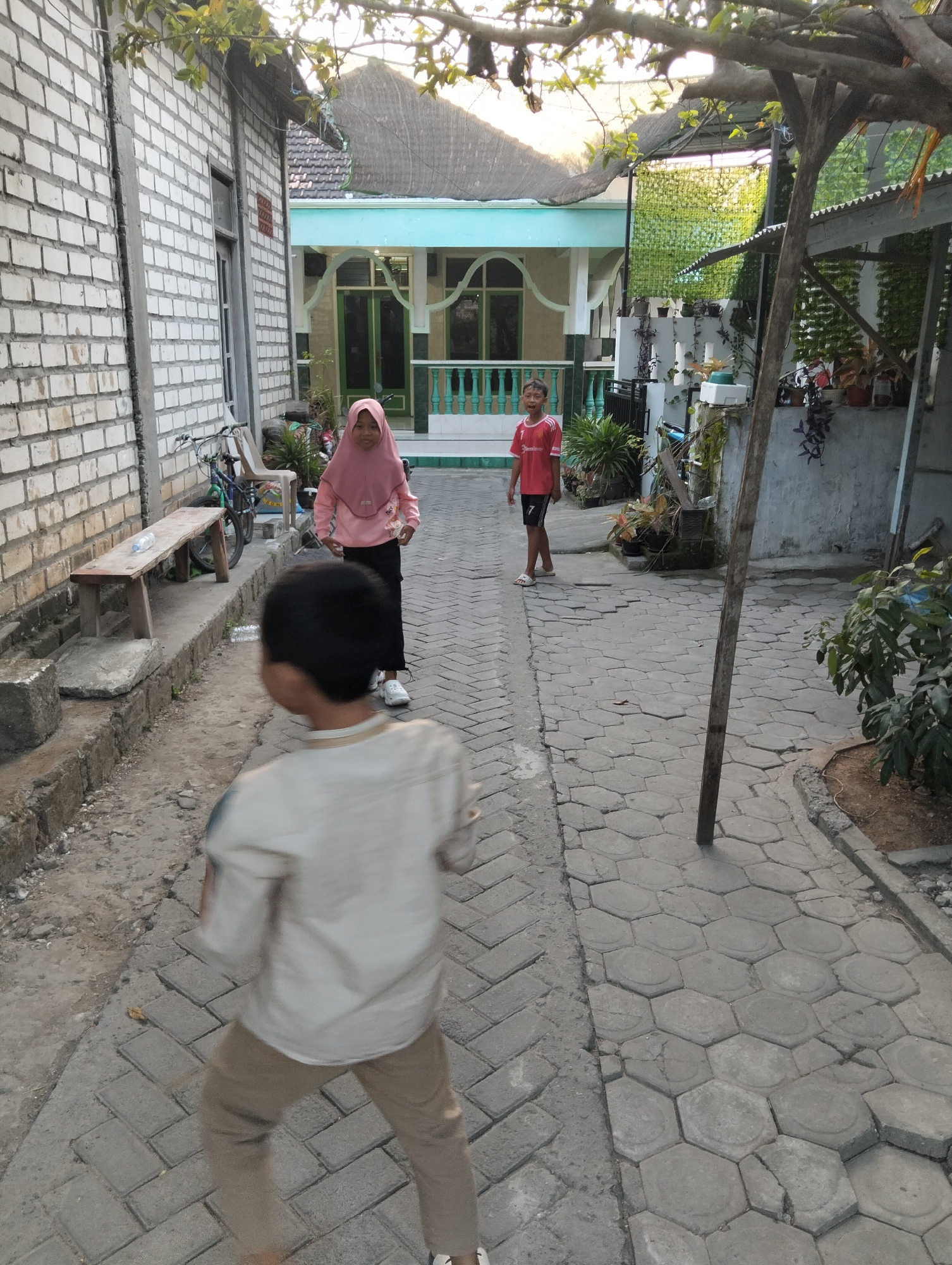 In Paciran, Indonesia, two children are visible on a paved walkway in front of residential buildings. A young boy, wearing a light-colored shirt and tan pants, is in the foreground, facing away from the camera and appearing to be in motion. Further down the walkway, a girl in a pink hijab and dark pants stands facing the camera. To her left, a boy in a red t-shirt and black shorts is also visible. The environment includes brick buildings with white paint, a wooden bench, bicycles, potted plants, and a wooden post supporting an overhead structure. The walkway is constructed of interlocking concrete pavers. The lighting suggests it is late afternoon.
