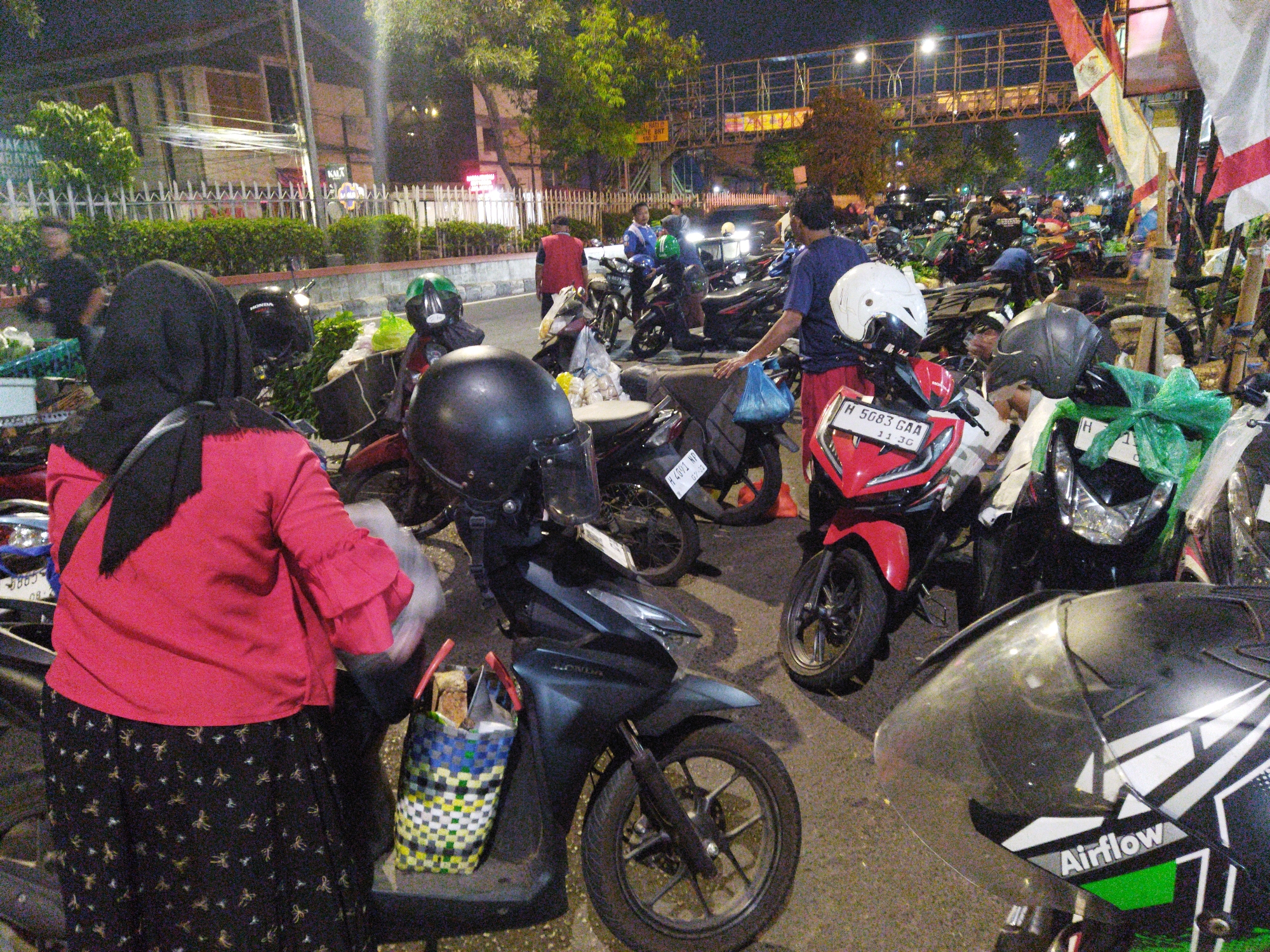 This is a nighttime photograph taken in Semarang, Indonesia, depicting a street scene likely in a busy area with many parked motorcycles. In the foreground, a person wearing a red shirt and a black headscarf is standing next to a dark-colored scooter. The scooter has a colorful woven basket attached to the front, filled with what appear to be bread or pastries. To the right of this scooter, another scooter, this one red, is visible, also with a helmet on its handlebars. Further in the background, numerous other motorcycles are parked. Some people are milling around, with one individual in a blue shirt and red shorts standing near a scooter, holding a blue plastic bag. Another person in a blue shirt and dark pants is visible further back. The environment appears to be an urban setting, possibly a market or a street where vendors are operating, as suggested by the basket of goods. There is an elevated structure in the background, possibly an overpass or part of a building. The lighting is artificial, typical of nighttime urban areas.
