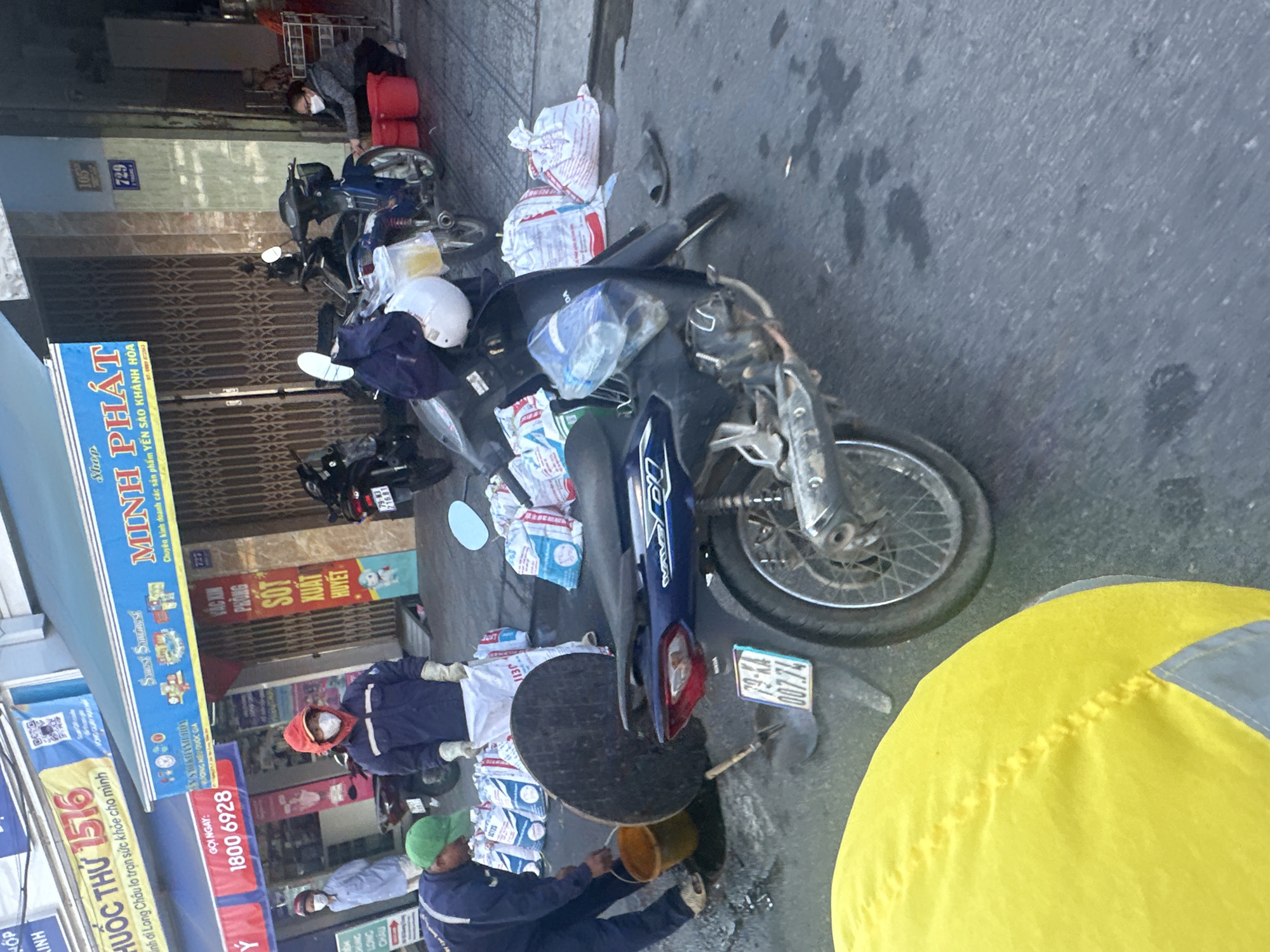 The image depicts a street scene in Bac Nha Trang Ward, Vietnam, where several individuals are engaged in what appears to be street cleaning or maintenance. A man in a blue uniform and green cap is actively pouring liquid from a yellow bucket onto the ground, near a large, dark circular object that could be a manhole cover. Beside him are several white bags, some with blue printing. Another person in a dark blue jacket and red hood, wearing a face mask and gloves, is holding a white bag and appears to be contributing to the activity. In the background, a woman in a grey jacket and face mask is crouching near red buckets. Several motorcycles are parked nearby. The environment is an outdoor urban setting with shopfronts visible in the background, one with a blue and yellow sign that reads "MINH PHÁT". The ground is wet in places, suggesting recent cleaning or a spill. The overall impression is one of a community effort or a work-in-progress on the street.
