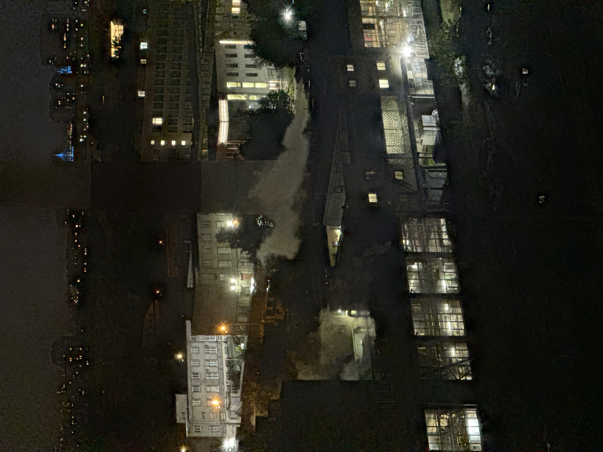 The image is a night-time aerial view of an industrial area in Sinyavino, Russia. Several multi-story buildings with numerous windows are visible. Some buildings appear to be factories or industrial complexes, with smoke or steam emitting from them. The overall scene suggests an active industrial site at night. The distant background shows a cityscape with lights from other buildings.