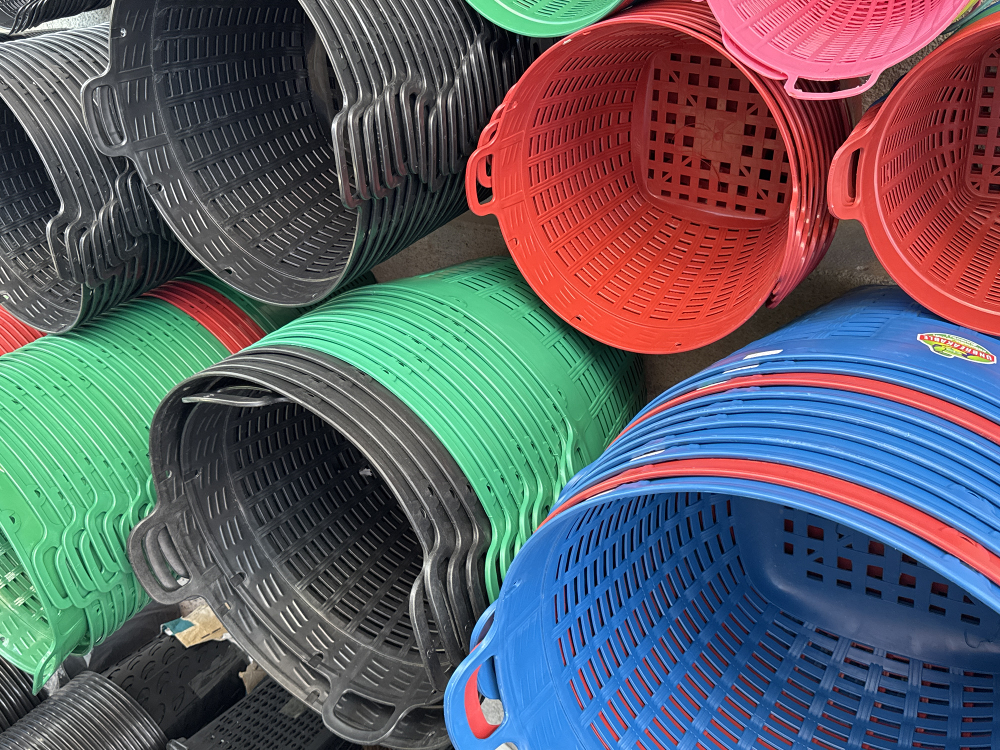 A close-up, high-angle shot captures a jumbled collection of stacked plastic baskets in various bright colors, including green, black, red, blue, and pink. The baskets are primarily utilitarian in design, featuring perforated sides and sturdy handles. Some of the baskets are nested inside one another, creating a sense of depth and volume. The lighting is natural, suggesting an outdoor setting or a well-lit indoor space. The overall impression is one of abundance and a marketplace or storage area where these baskets are readily available. The specific location in the city of Ranong, Thailand is not discernible from the image itself.