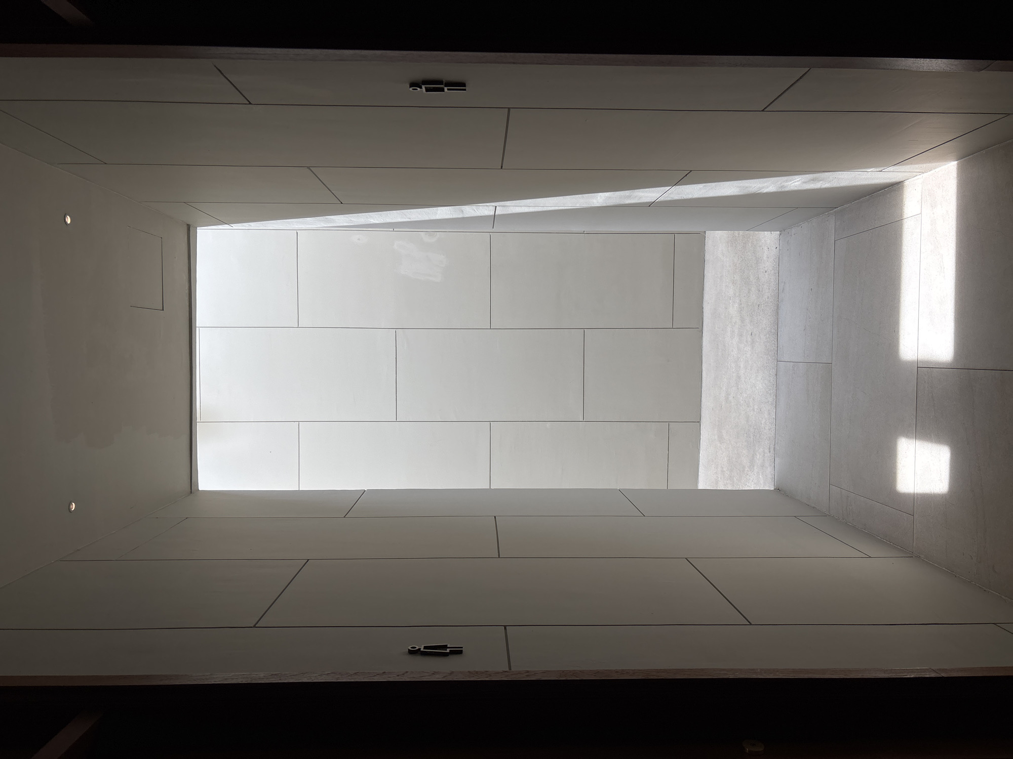 A low-angle, symmetrical shot depicts a rectangular interior space constructed from large, light-colored rectangular tiles or panels. The composition is dominated by converging lines leading to an opening at the far end, where light streams in. The side walls have subtle lighting elements. Two small, stylized human figure icons are affixed to the ceiling. The photograph is taken from a dark area, possibly an entrance, adding to the contrast in brightness. The scene appears to be a modern or minimalist architectural design. The location in Choeng Thale, Thailand is difficult to ascertain without additional context or identifying elements within the image.