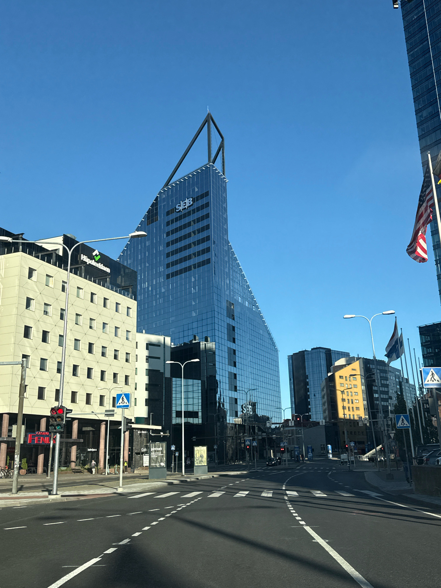 This is a street-level, eye-level, medium shot photograph taken in Tallinn, Estonia, on a clear, sunny day. The scene depicts a modern urban environment with several prominent buildings and a street in the foreground.
The central focus of the image is a tall, angular skyscraper with a glass facade that reflects the blue sky. The building has the letters "SEB" prominently displayed near its apex. To the left of this skyscraper is a lighter-colored, multi-story building with a grid-like pattern of windows. This building features signage that reads "Hüpoteeklaen" with a green logo. Further to the right, another tall skyscraper with a dark glass facade is visible, and American and Thai flags are flying on poles in front of it. In the distance, other buildings of varying heights and architectural styles are visible.
The foreground features a paved street with white dashed and solid lane markings. A pedestrian crossing is marked on the road. Traffic lights are visible, with some showing red signals. Streetlights and traffic signs, including a pedestrian crossing sign, are also present. A few individuals can be seen walking on the sidewalk or near the buildings. The overall impression is of a business district in a contemporary city.