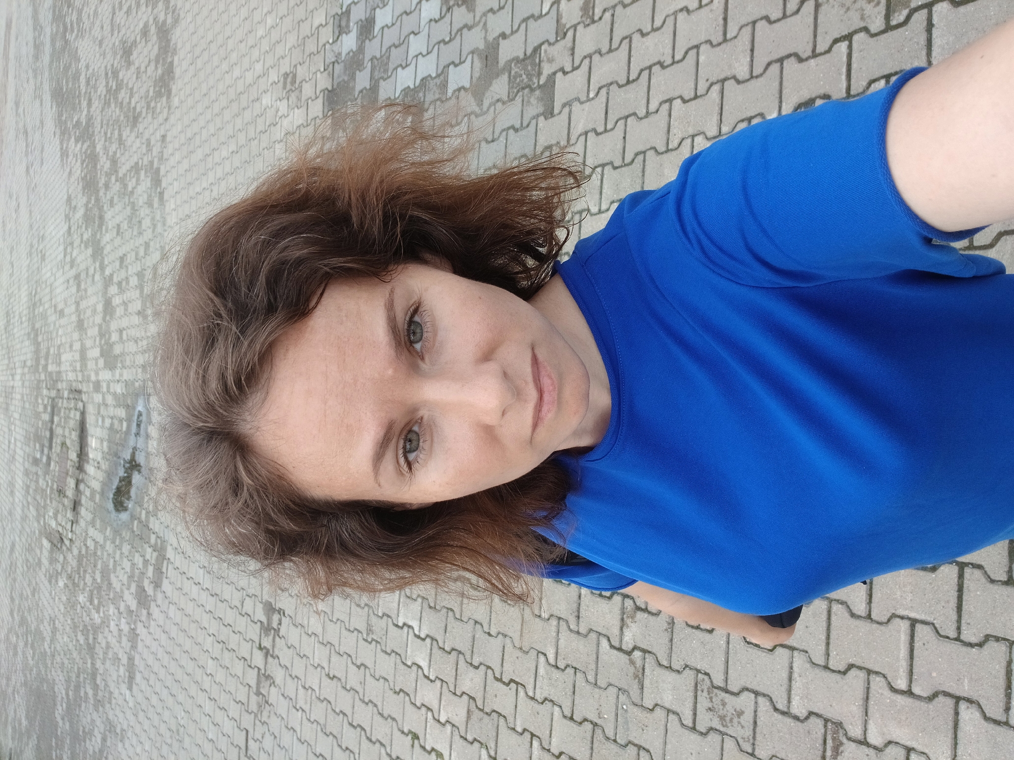 A woman with brown, curly hair is captured in a high-angle, selfie-style shot. She is wearing a bright blue t-shirt and looking directly at the camera with a neutral expression. Her face is slightly tilted, and the focus is on her facial features, including her eyes and freckled skin. The background consists of a paved area made of interlocking concrete blocks, appearing damp in places, suggesting an outdoor urban environment. The lighting indicates it is daytime. The scene is devoid of any other individuals or prominent objects.