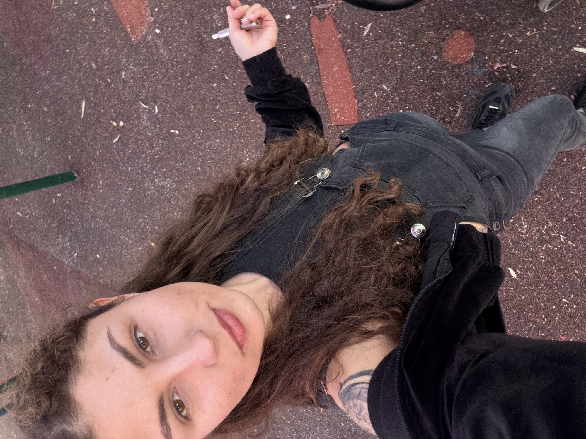 This is a high-angle, full-body portrait photograph of a young person, likely female, with long, curly brown hair, lying on an outdoor ground surface. The subject is looking directly up at the camera with brown eyes and a neutral to slightly pleasant expression. She has a small nose ring on her left nostril and some freckles on her face.
She is dressed in dark attire, including a black long-sleeved top, a dark gray or black denim jacket or vest with distinctive silver buckles/straps on the chest, and dark gray jeans. Her left arm reveals a visible tattoo peeking from under her sleeve. She wears black sneakers on her feet. In her raised right hand, she holds a silver-colored cylindrical object, resembling a vape pen or e-cigarette.
The setting appears to be an outdoor area in Kiryat Motzkin, Israel, with the person resting on a textured ground surface that is predominantly dark reddish-brown with lighter speckles, possibly a rubberized floor or asphalt. Scattered debris and brighter reddish-brown patches are visible on the ground. A narrow green line runs along the left edge of the frame on the ground. The lighting suggests it is daytime. There is no visible text in the image.