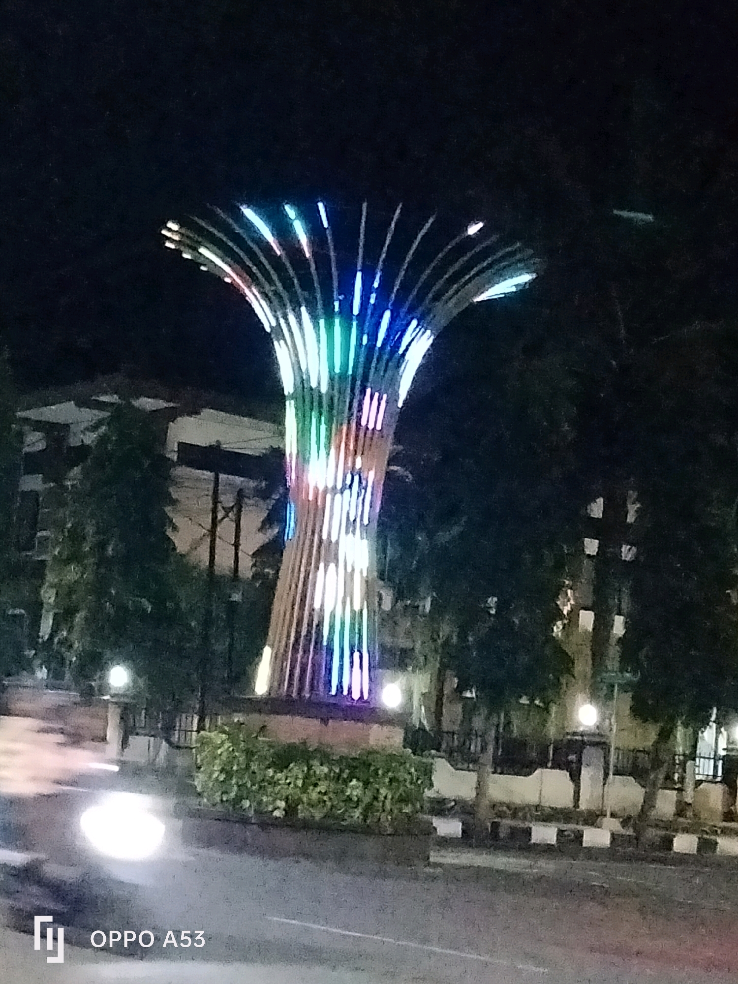 The image depicts a modern, illuminated sculpture at night, likely a landmark in the city of Trenggalek, Indonesia. The sculpture, characterized by its tall, fluted design, is adorned with a vibrant array of colorful LED lights, primarily in shades of blue, green, and pink. It stands amidst a landscaped area with green bushes at its base, bordered by a checkered pattern of white and black concrete barriers. Behind the sculpture, buildings and trees are visible, dimly lit against the dark night sky. A street runs in the foreground, with a bright, diffused light reflecting on its surface, possibly from vehicle headlights or street lamps. The watermarked text "OPPO A53" in the bottom left corner indicates the device used to capture the photograph.