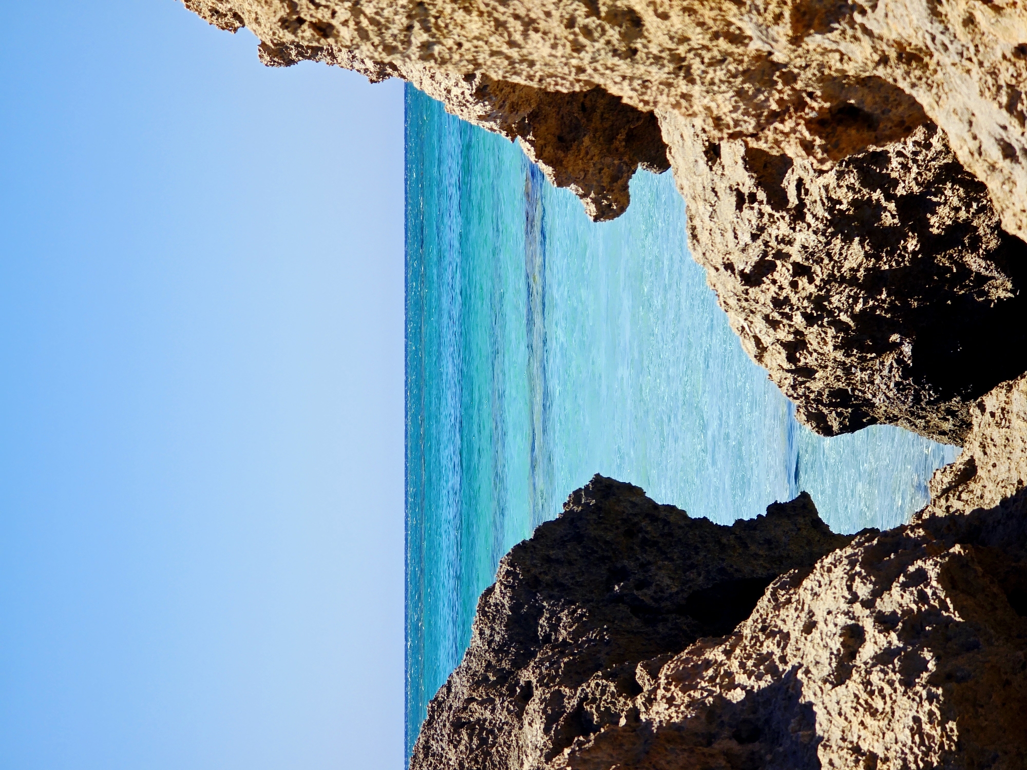 The image depicts a coastal scene with clear turquoise water and a bright blue sky. In the foreground, two large, textured, rocky outcrops frame a view of the sea. The rocks are a light brown or tan color with rough surfaces and visible pores. The sea is calm with a gradient of blue and green hues, indicating shallow, clear water. The horizon line is sharp, separating the water from the expansive, cloudless sky. The location is described as Unknown, Greece. There are no people or discernible actions occurring in the image.