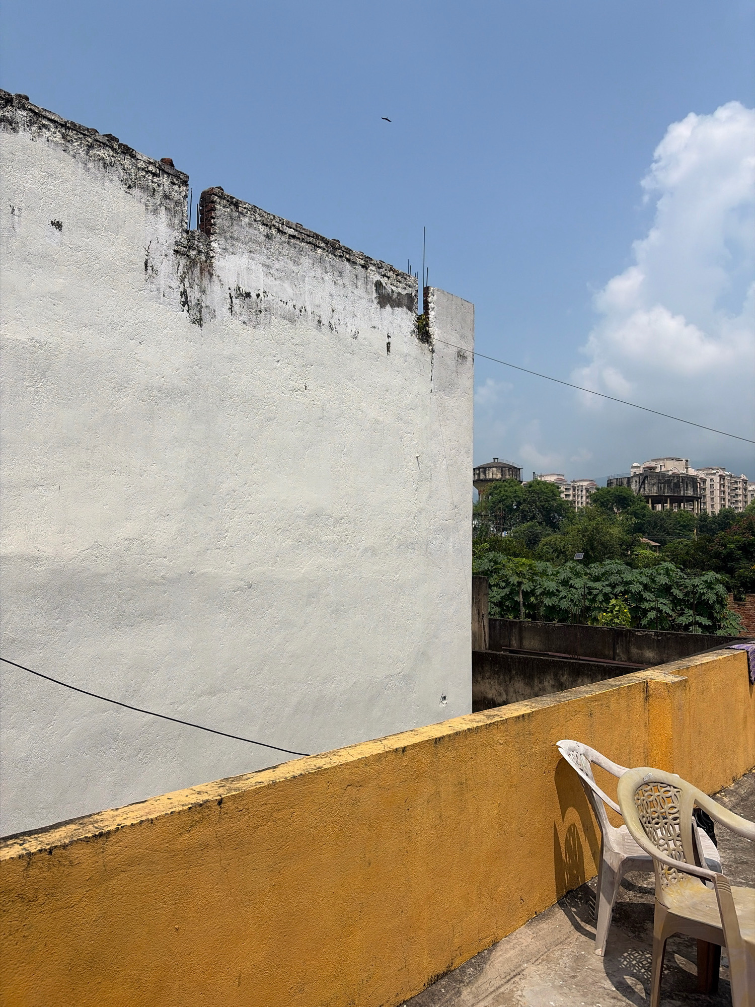 The image depicts a rooftop scene in Jamshedpur, India, on a clear day. In the foreground, a textured, yellow-painted wall runs horizontally across the frame. Two plastic chairs, one white and one beige with intricate cutouts on the back, are placed on the concrete rooftop to the right. A shadow of one of the chairs is cast on the yellow wall.
A large, rough-textured white wall dominates the left and middle ground of the image. It appears weathered and shows signs of damage along its top edge, with exposed brickwork and some vegetation growing out of it. A black wire is visible stretched horizontally across the white wall.
In the background, a dense canopy of green trees separates the foreground from a cityscape. Several multi-story buildings, including some with water towers on top, are visible beyond the trees. The sky is a clear blue with scattered white clouds. A small bird is silhouetted against the sky in the upper left portion of the image.