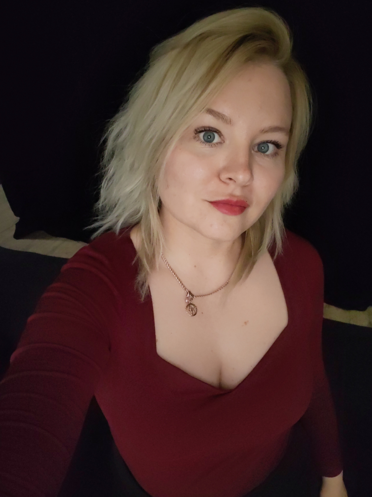 A young woman with light blonde, shoulder-length hair is featured in a medium close-up selfie. Her striking light blue eyes gaze directly at the camera, conveying a neutral yet composed expression. She is wearing a warm red lipstick, and a small beauty mark is visible near the left corner of her mouth.
She is dressed in a dark red or maroon long-sleeved top with a wide, squared neckline. A delicate rose gold-toned chain necklace with a small, circular pendant, possibly depicting a detailed emblem or figure, adorns her neck and chest.
The background is largely dark, suggesting an indoor setting with subdued lighting or a plain dark backdrop. Faint outlines of lighter-colored objects or fabric can be seen in the lower left and right areas of the frame, hinting at furniture or decor. The overall lighting illuminates her face and upper body, creating a soft contrast against the dark surroundings. The image captures a posed moment, likely a self-portrait. No text is visible in the photograph. This image is contextualized as being from Izhevsk, Russia.