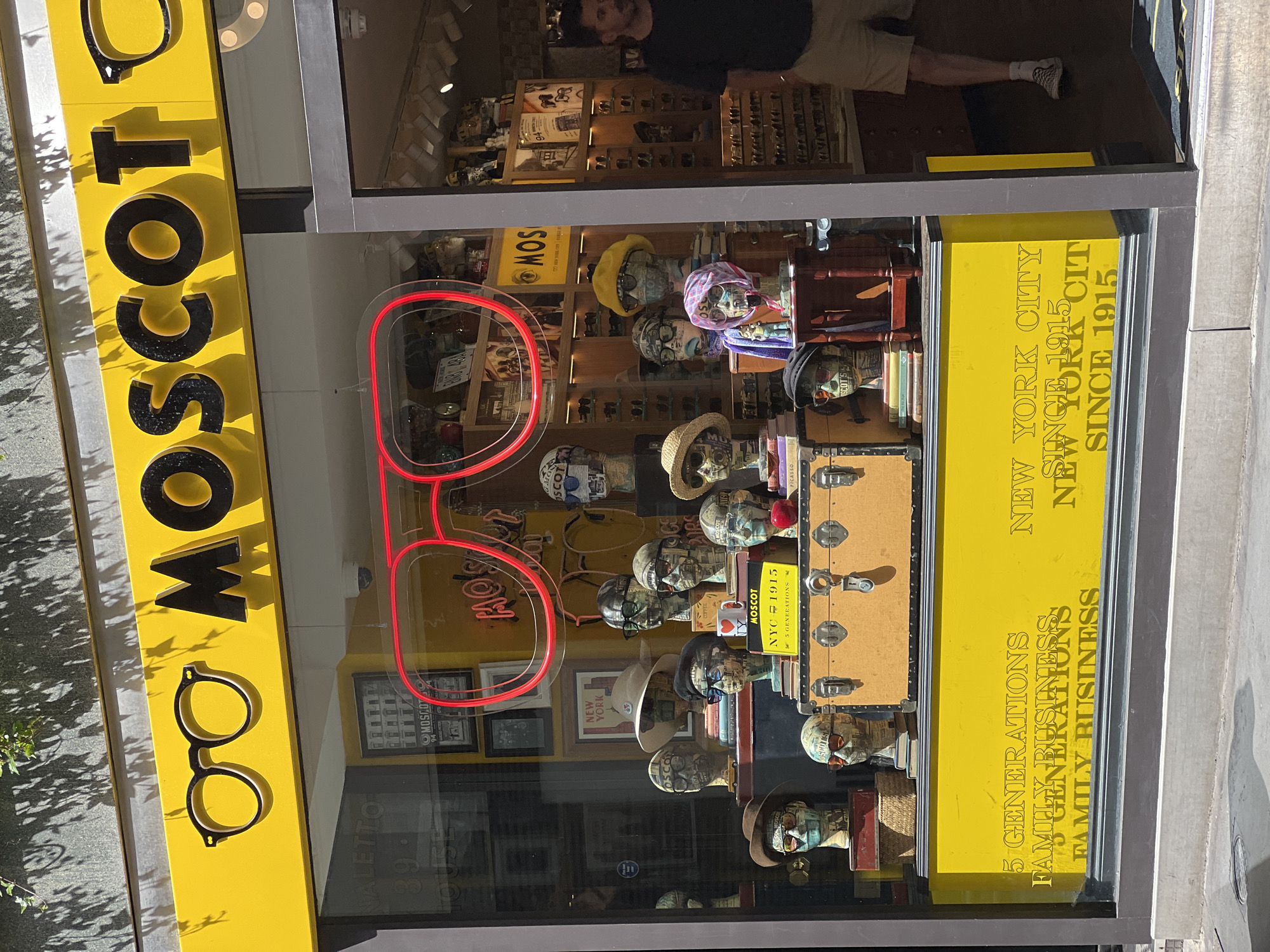 This is a photograph of the Moscot eyewear store. The exterior of the store is visible. The sign is yellow with the company name written in black letters, with a picture of glasses. The window displays rows of mannequins wearing glasses. A neon sign of the brand name is visible on the glass. The window also displays details about the company, including "New York City since 1915" and "5 Generations Family Business". A man is visible in the reflection on the top part of the window.
