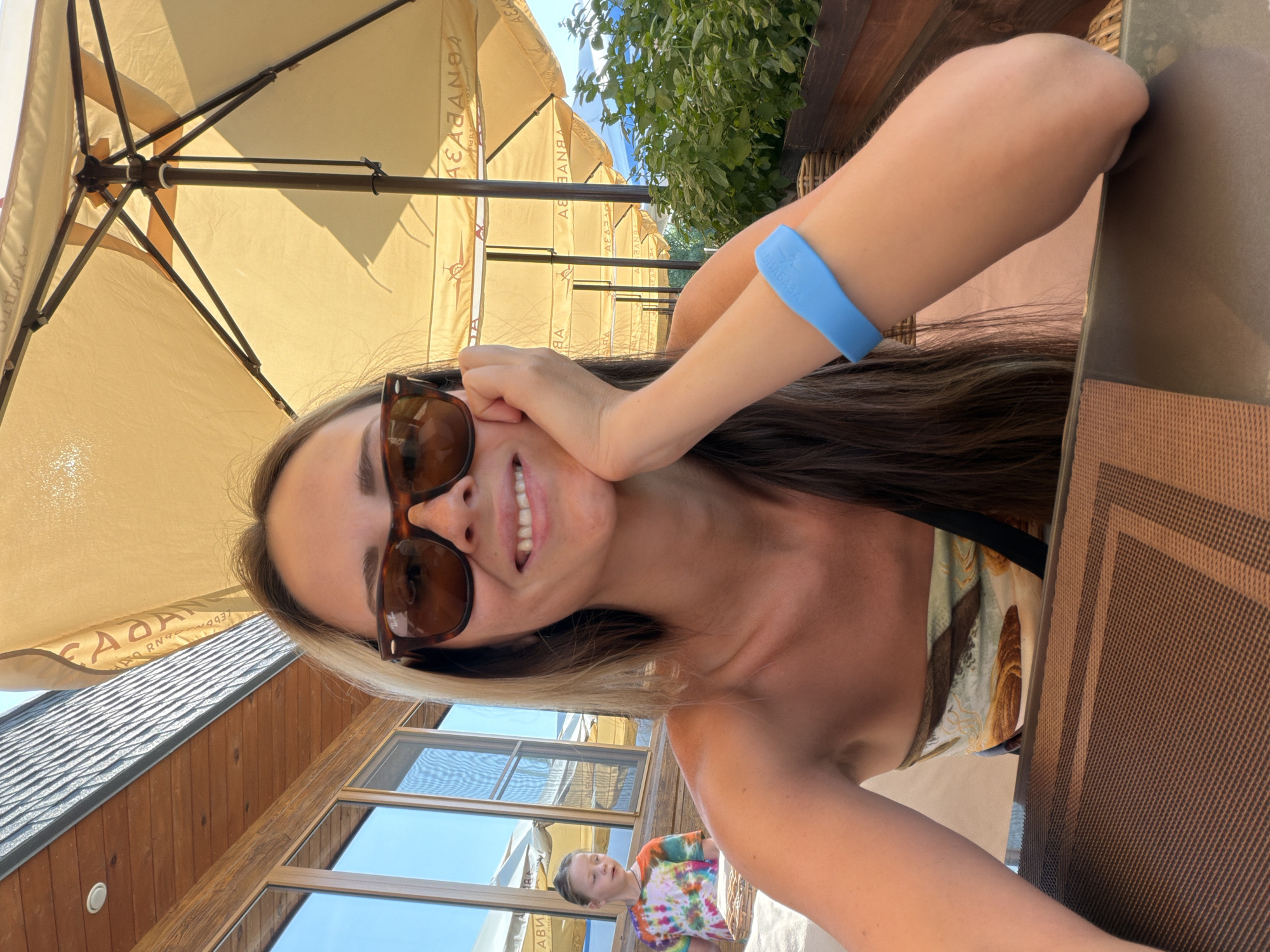 A young woman with long, light brown hair and sunglasses is seated at a table outdoors, likely at a cafe or restaurant, evidenced by the table setting and umbrellas. She is resting her cheek on her hand and smiling. She wears a strapless top with a pattern that appears to be shells and waves. A blue silicone wristband is visible on her right arm. In the background, a young girl with blonde hair, wearing a tie-dye t-shirt, is visible through a window or glass door. The setting appears to be a sunny day, with wooden building elements and lush greenery visible. The specific location in Kamyshin, Russia is not discernible from the image itself, but the presence of Cyrillic script on the umbrellas suggests a Russian-speaking country.