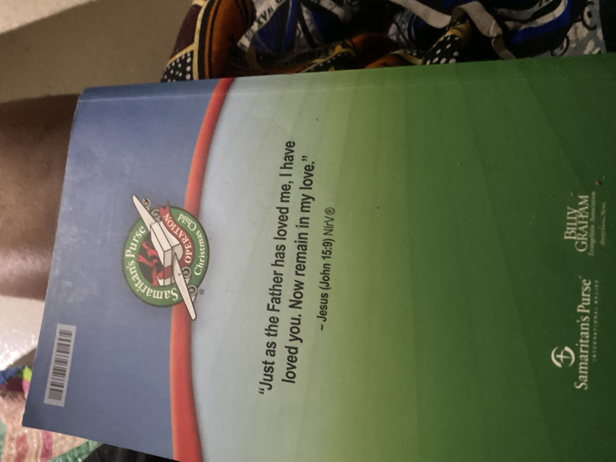 The image shows the back cover of a book. At the top left, there is a barcode. A circular logo with "Samaritan's Purse Operation Christmas Child" is visible in the upper middle of the cover. Below the logo, a quote is written in white text: "Just as the Father has loved me, I have loved you. Now remain in my love." Below the quote, it is attributed to "- Jesus (John 15:9) NIrV®". In the bottom left corner, the Samaritan's Purse logo with the text "SAMARITAN'S PURSE" is visible. To the right of this, partially visible, is the "BILLY GRAHAM EVANGELISTIC ASSOCIATION" logo. The background of the book cover transitions from blue at the top to green at the bottom. The book is lying on a patterned fabric. There is no information in the image to suggest a location in Jauro Yinu, Nigeria.