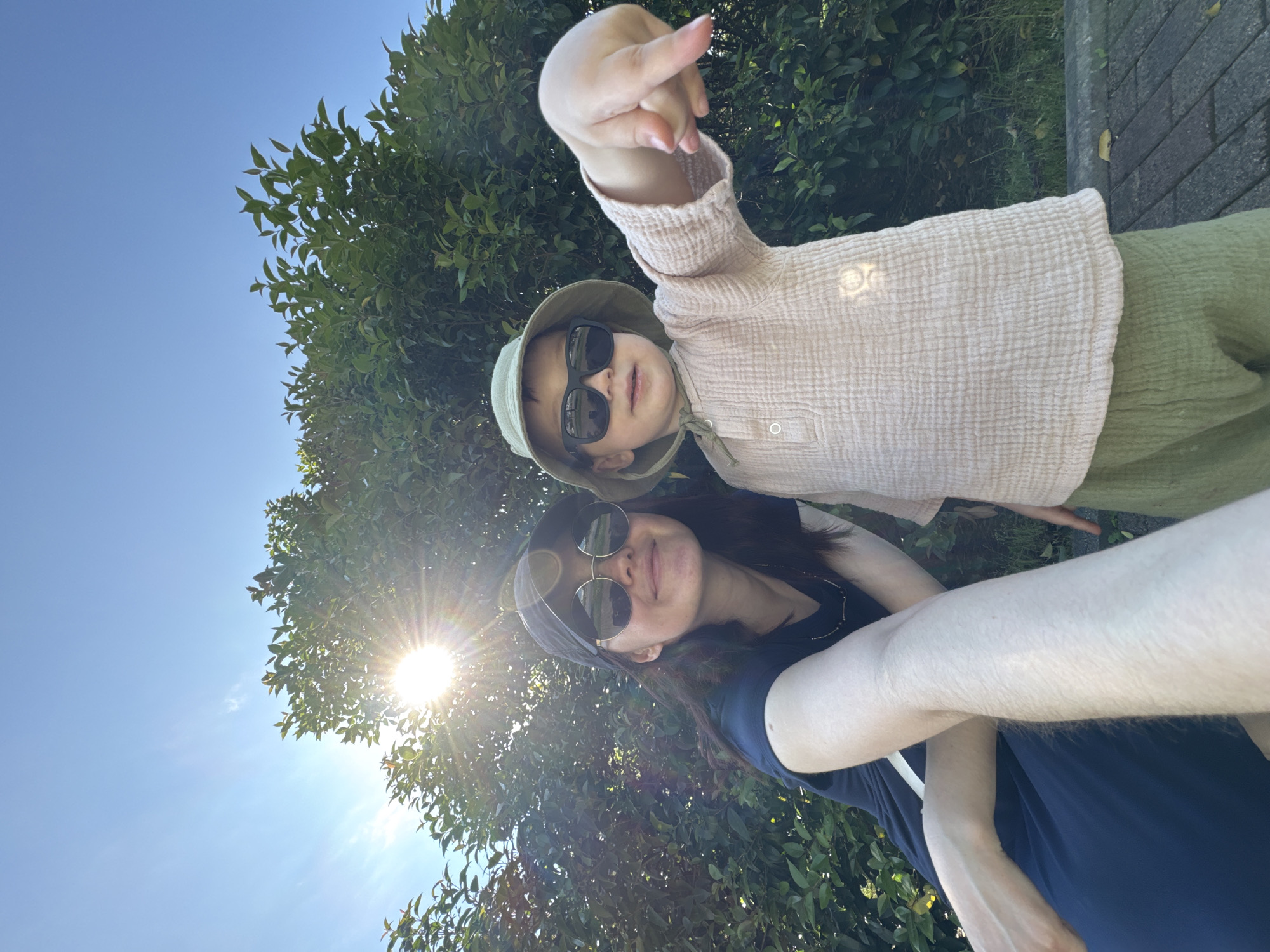 A low-angle selfie captures a woman and a child outdoors, with sunlight filtering through trees overhead. The child, positioned above the woman, wears sunglasses, a bucket hat, a light pink textured top, and green shorts. The child's right arm is raised, with fingers extended forward. The woman, also wearing sunglasses, has dark hair and a dark-colored top. The background consists of green foliage, a clear blue sky, and a section of paved ground. The location is Sirius, Russia.