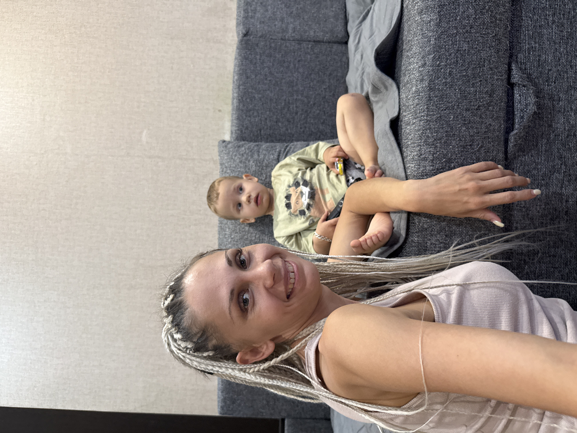 A woman with blonde braided hair and a light pink tank top is in the foreground, smiling at the camera. Behind her, a young boy with short brown hair is sitting on a grey sofa, looking to his left. He is wearing a light green t-shirt with a lion graphic and patterned shorts. A grey blanket is draped over the sofa. The environment appears to be a domestic setting, possibly an apartment in Rusanovo, Russia, with a textured wall in the background.