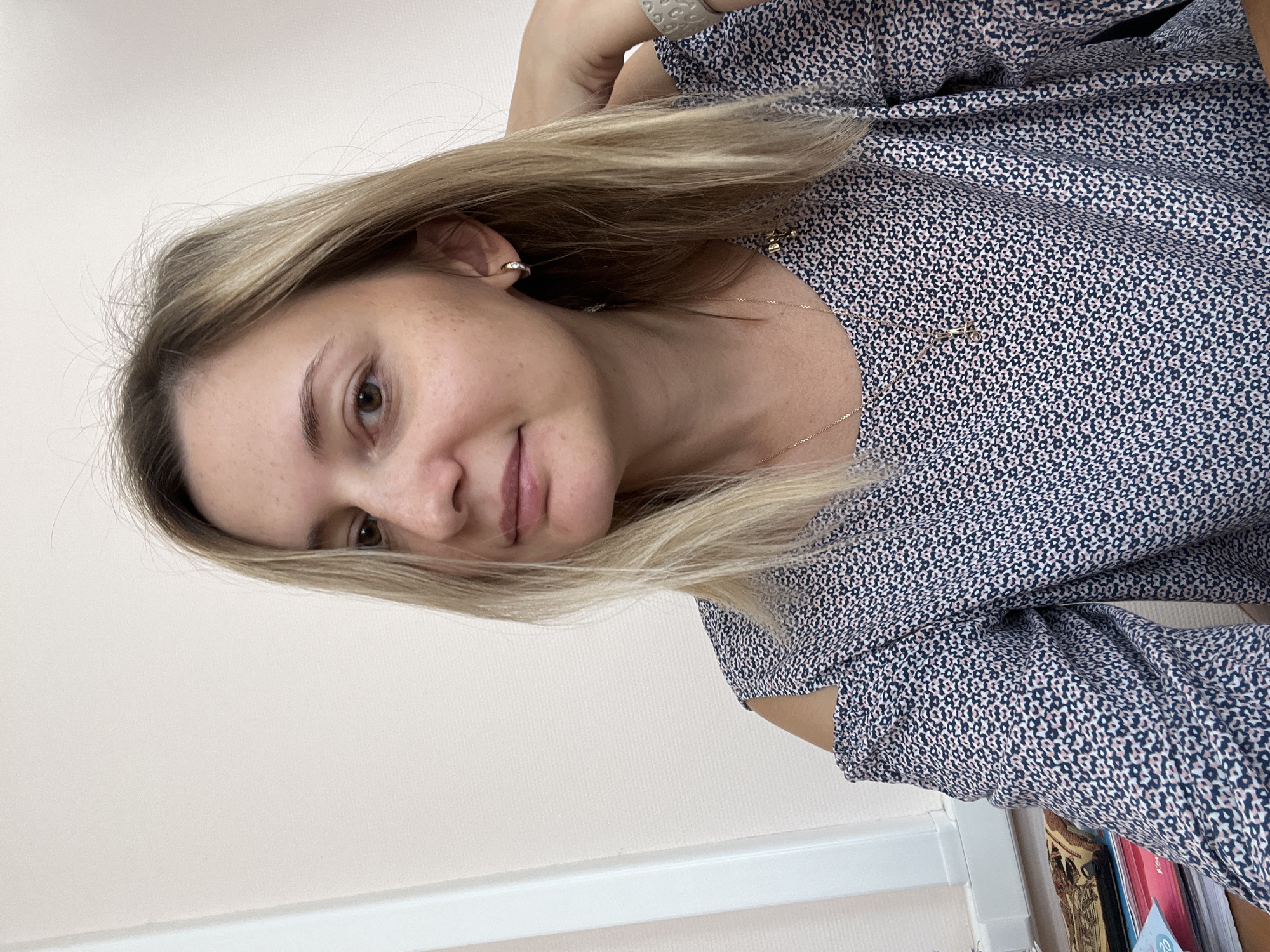 A selfie of a fair-skinned Caucasian woman with blonde hair, light brown eyes, and freckles is shown. She is wearing a blue and pink patterned blouse with cold shoulders and a silver hoop earring in her left ear. A silver necklace with a cross pendant is visible around her neck. She has a watch on her right wrist. The woman is leaning her head on her hand, and her hair is partially obscuring her face. The background is a light-colored wall and a window with vertical blinds on the left side of the image. On the desk in front of her, there are stacks of papers and a book. The image appears to have been taken indoors, likely in an office or study. There is no discernible information to suggest this scene is located in the city of Volgograd, Russia.