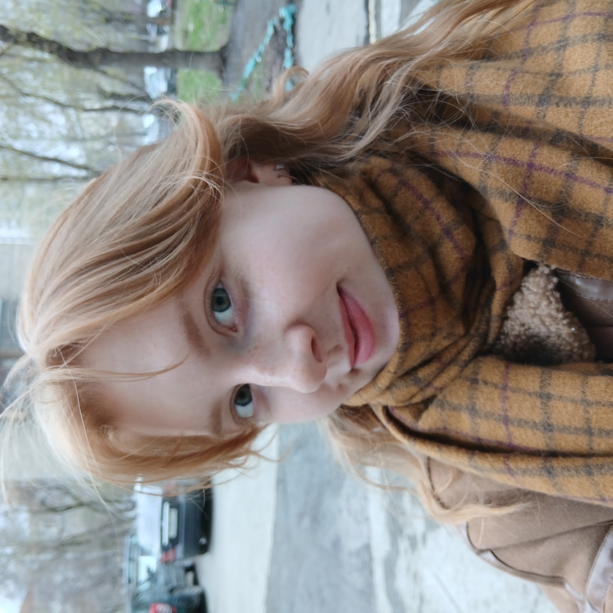 A close-up portrait depicts a young woman with fair skin, freckles, and light reddish-blonde hair. Her hair is styled loosely, framing her face. She has light-colored eyes and is wearing lipstick or a lip tint. She is wearing a tan jacket with a plaid scarf wrapped around her neck. The background shows urban elements. The location of the scene is the city of Yekaterinburg, Russia. A car is parked behind her. Bare trees suggest an outdoor environment.