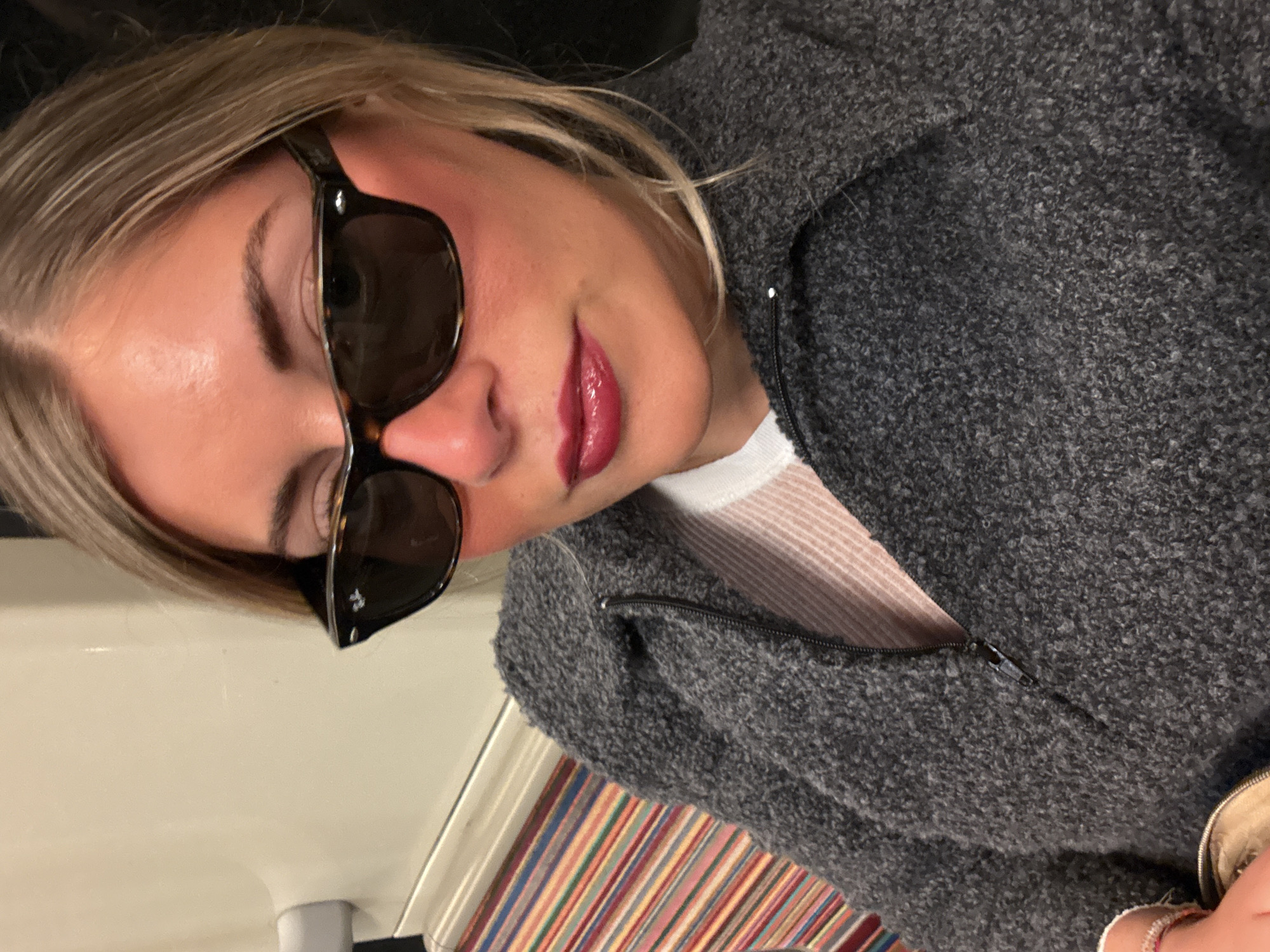 A woman with blonde hair and dark sunglasses is wearing a gray, fleecy zip-up jacket. She appears to be seated inside a carriage in the Paris metro. The colorful striped seating is visible in the background.