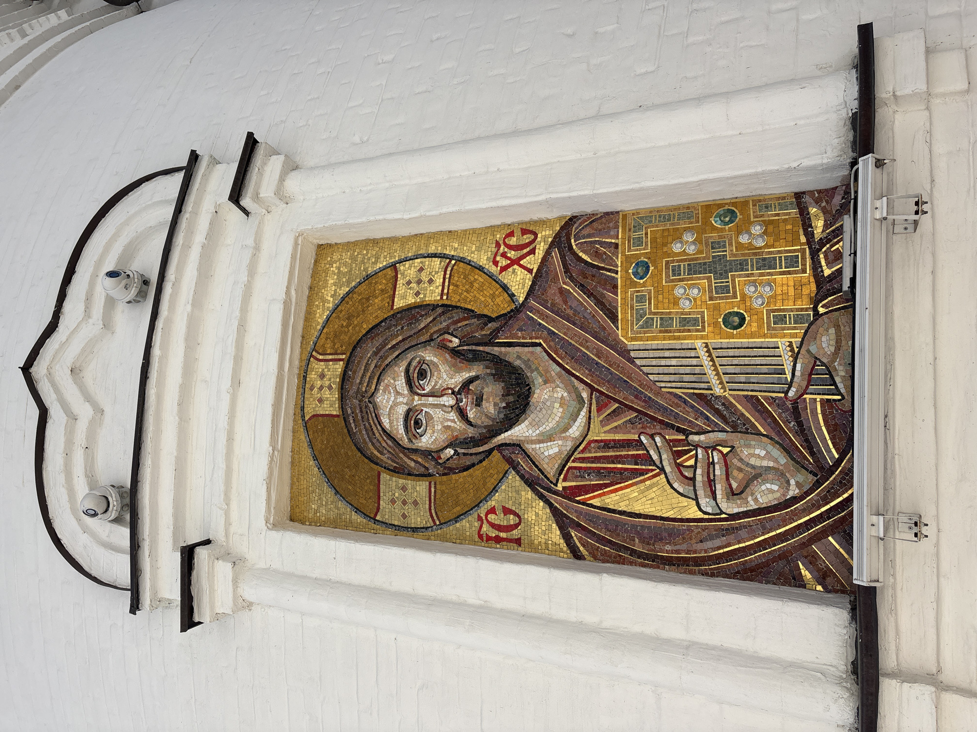 The image shows a mosaic of Jesus Christ mounted on a white wall. The mosaic features Jesus' head, halo, and part of his upper body with his hand raised in blessing. The mosaic is primarily composed of gold, brown, and red tiles. There are red Greek letters present near his head.
The mosaic is framed by an ornate white border with black metal detailing. A security camera is visible near the border. A metal pipe with mounting hardware is affixed to the wall near the right side of the mosaic.
Based on the information available, it is not possible to determine the specific location of this mosaic in Ulyanovsk, Russia, or whether it is even in Ulyanovsk.