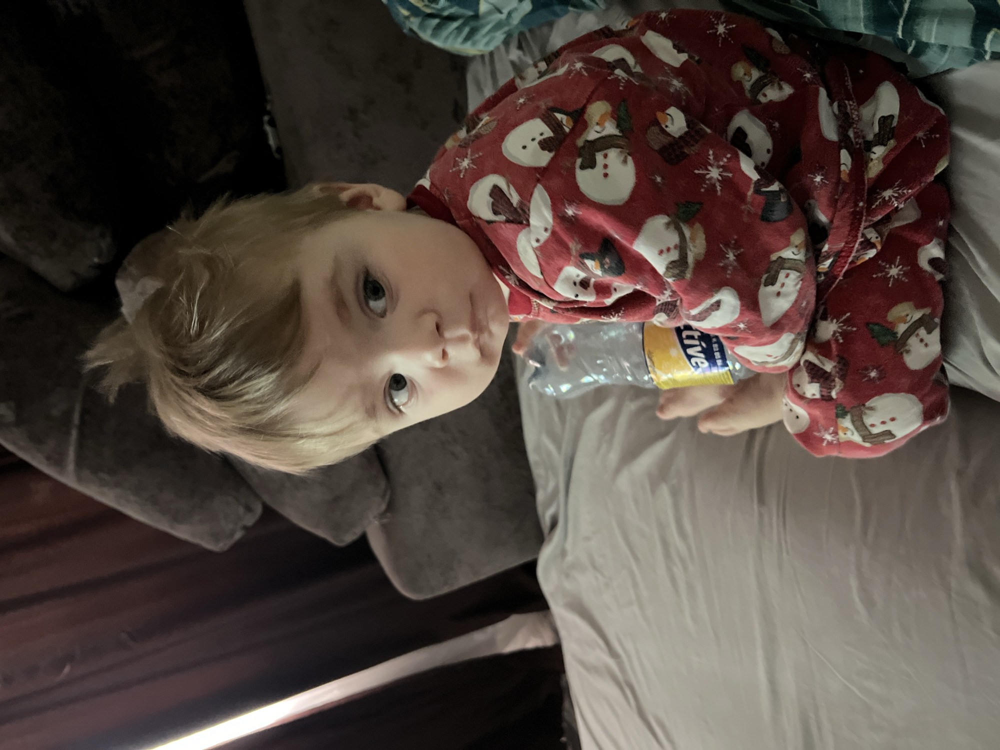 A young child with blonde hair, wearing red pajamas with snowman patterns, is lying on a gray bed. A water bottle sits near the child. Part of a gray sofa is visible to the left.