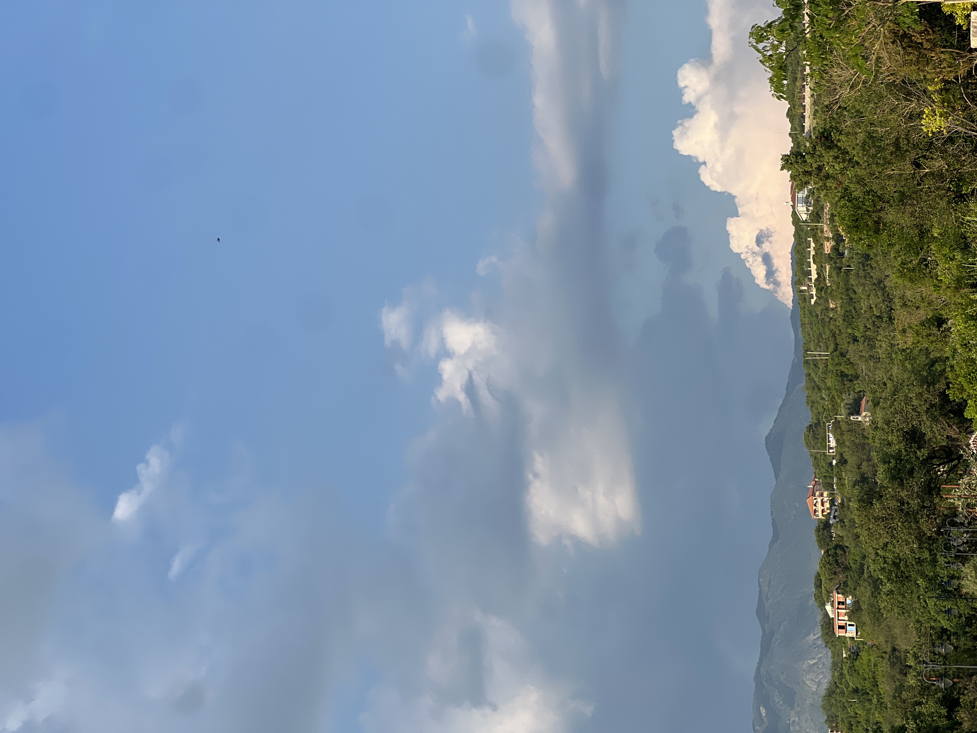 The image presents a view in Kovačko Polje, Montenegro. Dominating the right side of the frame are green, lush mountainsides dotted with residential buildings. The mountains fade into the background, suggesting distance. In the sky, expansive formations of clouds are visible, ranging from light and fluffy to dark and stormy. There is a small bird flying in the clear sky.