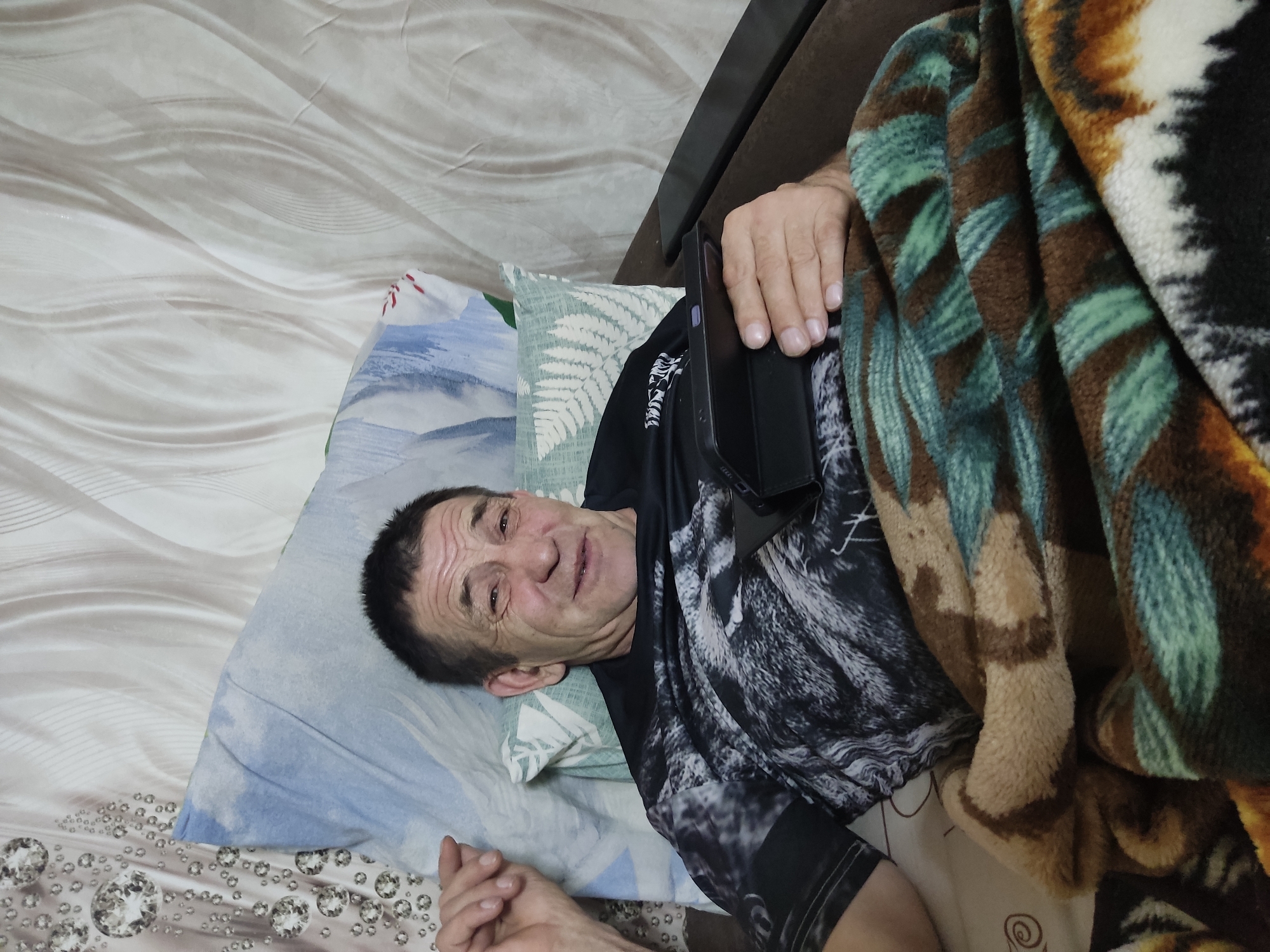 A man is lying in bed, covered by a blanket, and holding a tablet. The background shows a wall with a wavy pattern and a pillow with a blue and white design. The man is wearing a black t-shirt with a wolf design on it. He appears to be smiling and looking at the camera. The setting appears to be a bedroom. The image does not contain any information that would allow for the determination of a specific location within Noginsk, Russia.