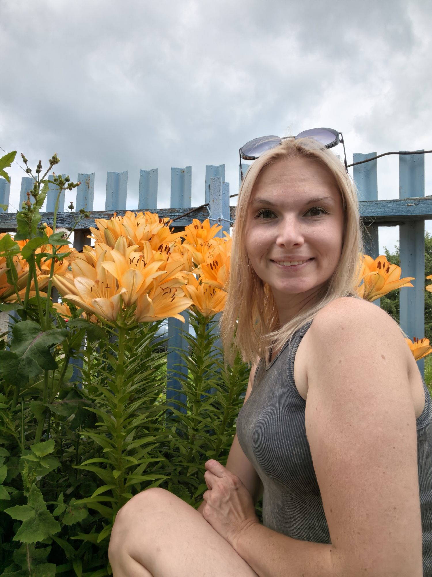 A full-shot, outdoor, selfie-style photograph features a blonde woman in a gray tank top and denim shorts. She is seated in front of a patch of vibrant orange lilies and a pale blue wooden fence. Sunglasses are perched on top of her head. The background consists of an overcast, cloudy sky. The overall impression is that of a casual moment captured in a garden or yard.
