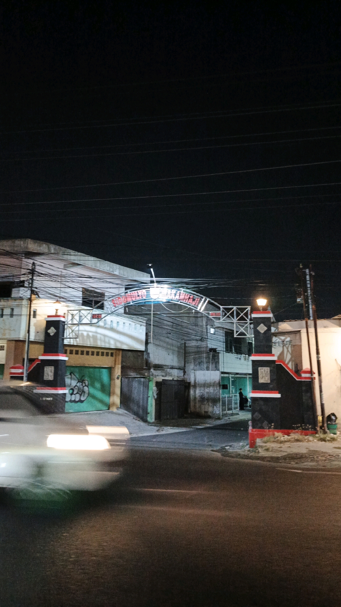 The image depicts an outdoor scene at night, likely in Pabelan, Indonesia. In the foreground, a blurred car is driving past, its headlights creating streaks of light on the wet asphalt road. The road curves slightly to the right.
The middle ground features an entrance or gate. Two pillars, one on each side, frame the entrance. These pillars are painted black with red and white accents. The left pillar has a plaque affixed to it. Behind the pillars, an arched structure spans the road. This arch is illuminated and appears to have text on it, though the words are difficult to decipher. A building with a light-colored facade and numerous electrical wires overhead is visible behind the arch. To the left of the arch, a green garage door with graffiti is partially visible.
In the background, the sky is dark, suggesting it is nighttime. The environment appears to be urban, with buildings and infrastructure like utility poles and overhead wires present. The overall atmosphere is dimly lit, with artificial lighting from the arch, the pillars, and the car's headlights illuminating the scene.
