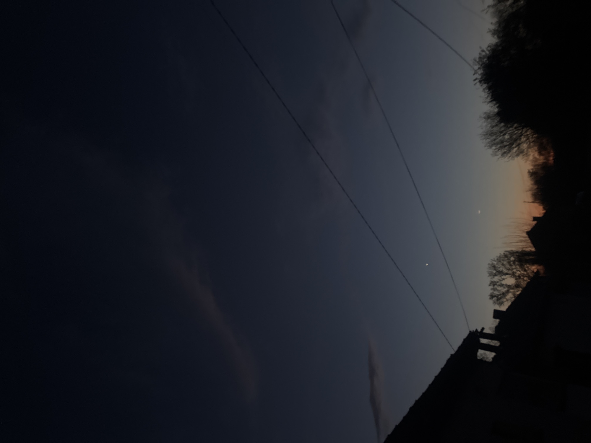 The image depicts a dusk or dawn scene in what appears to be a residential area. The sky transitions from a deep blue on the left to a lighter gradient on the right, with hints of orange near the horizon. Several power lines are visible crossing the frame diagonally. On the right side of the image, silhouetted rooftops and trees are present. A small, crescent moon is visible in the sky. The location is Lehchevo, Bulgaria, as specified.