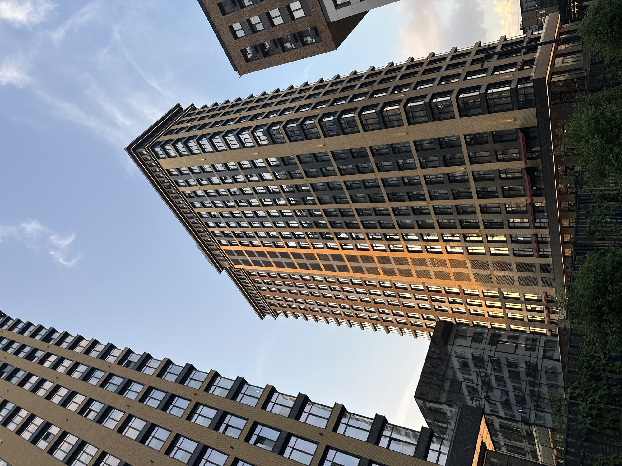 The image is an upward view of several modern buildings. The central building is a tall, rectangular structure with multiple stories. It has a patterned facade, alternating between beige and dark gray, with rows of windows on each floor. Sunlight is reflected on the central facade, creating a gradient of light. Other buildings surround the central structure, with one in the upper left and one in the lower left, both exhibiting similar modern architectural styles. In the right-hand side of the image there is greenery. The sky is visible between the buildings, with light blue hues and wispy clouds. The location is Khimki, Russia.
