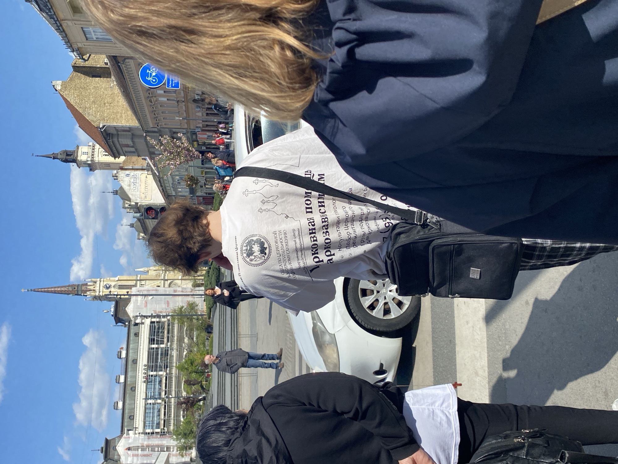 Here's a detailed description of the image:
The image captures a street scene in Novi Sad, Serbia, featuring several people and buildings. In the foreground, parts of three individuals are visible. A person with blonde hair is partially obstructing the view at the top. To their right stands a person with brown hair wearing a white T-shirt with Cyrillic text and a logo. The T-shirt design seems to relate to the Russian Orthodox Church and assistance to drug addicts. This person is also wearing a black shoulder bag. Below them, another person with dark hair is partially visible, dressed in a black jacket over a white shirt.
In the background, there's a white car, and a few people are walking along the street. A prominent building with a tall steeple and clock is visible. This is the Name of Mary Church, a major landmark in Novi Sad. Other buildings with varied architectural styles are present, adding to the urban landscape.
A blue bicycle lane sign is visible above one of the buildings. The sky is clear and blue, indicating a sunny day. The scene appears to be captured at ground level, and the perspective is slightly tilted.