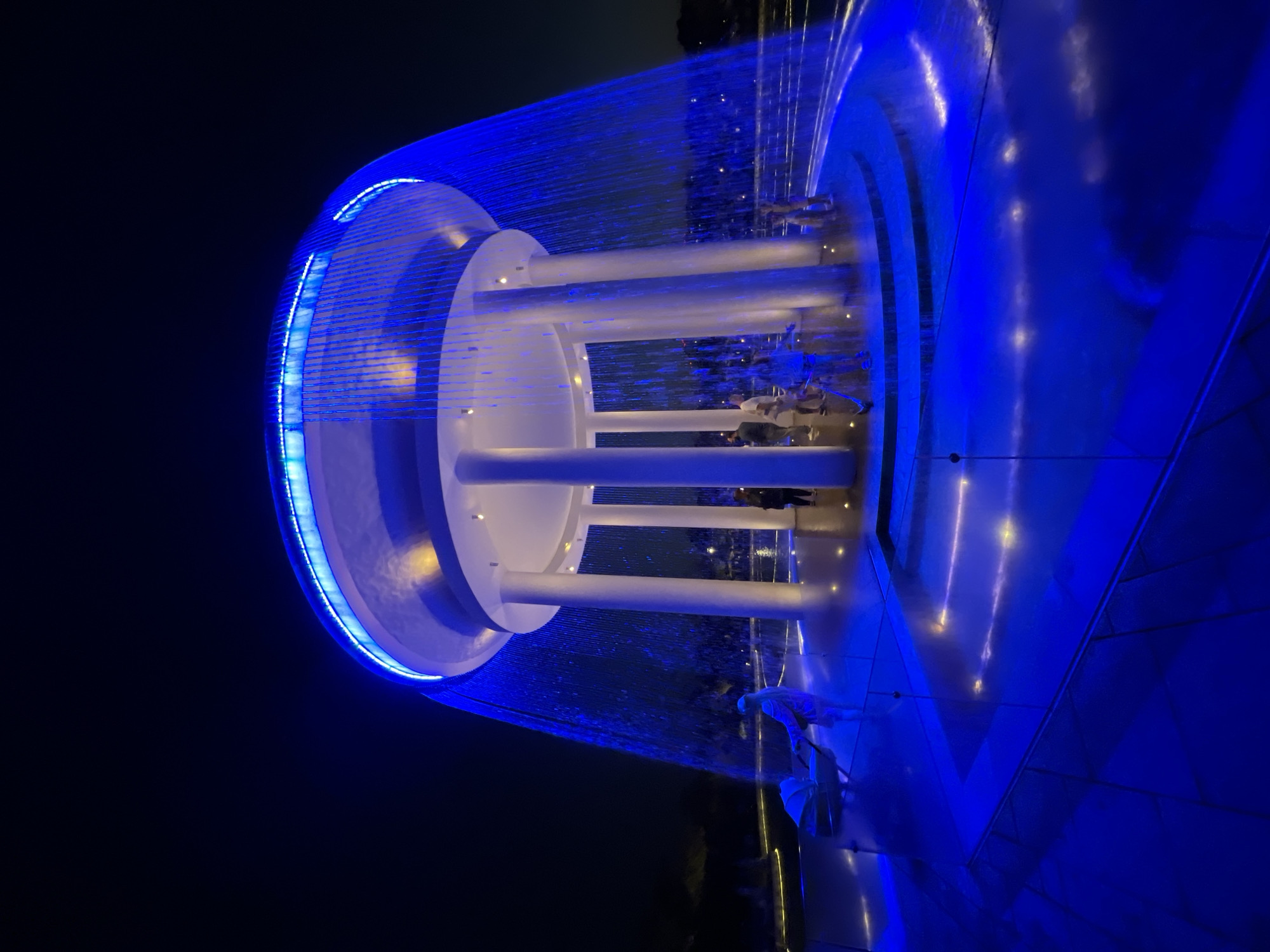 The image displays a modern water feature at night, illuminated by vibrant blue lights. The central structure of the fountain is a white, cylindrical tiered design with several large, white pillars supporting the upper levels. A curtain of water cascades down from the upper sections, creating a shimmering effect against the dark background. The water is caught in a large, circular pool or basin, the surface of which reflects the blue illumination, creating streaks of light.
Several people are visible in the scene, standing around the fountain and within its illuminated perimeter. They appear to be casually observing or interacting with the water feature. Their attire suggests a normal evening outing.
The setting is dark, indicating it is nighttime. The location appears to be an urban or park-like area, possibly a public square or a modern entertainment complex, given the sophisticated design of the fountain. The overall mood is serene and visually striking, characterized by the interplay of light, water, and modern architecture. There is no visible text in the image. The context provided indicates the location is Krasnodar, Russia.