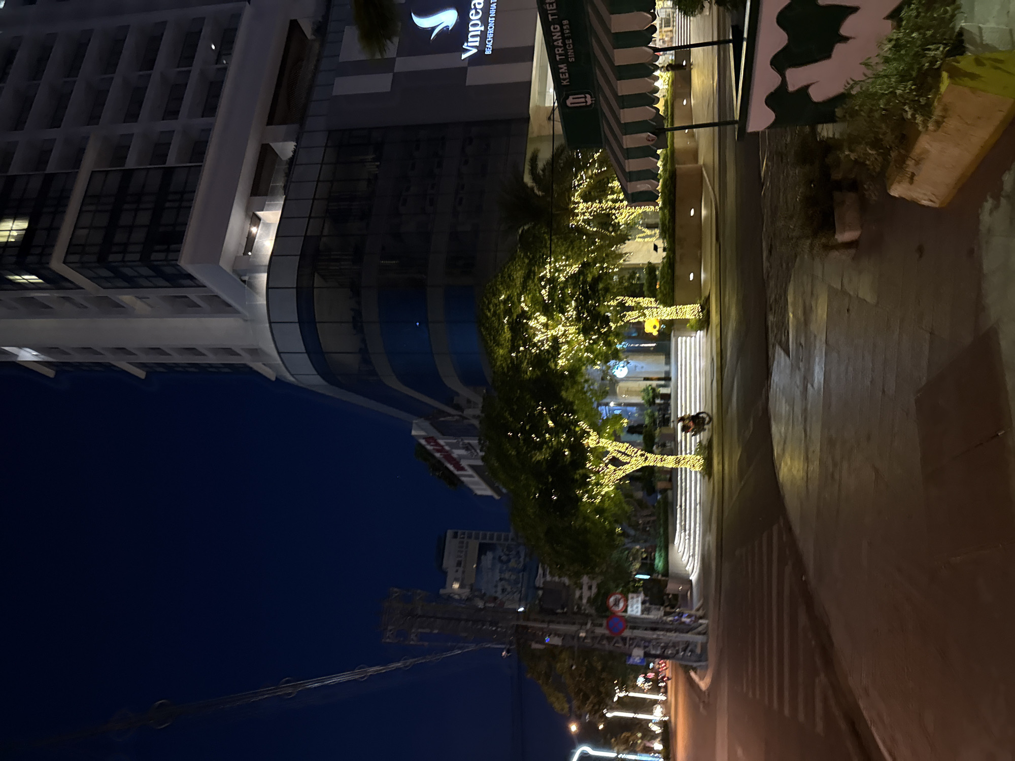 In the city of Nha Trang, Vietnam, a night scene unfolds with a modern high-rise building on the right, featuring the "Vinpearl Beachfront Nha Trang" logo. Adjacent to it, a row of trees are elaborately decorated with bright yellow string lights, creating a festive atmosphere. These illuminated trees stand near a set of white, illuminated stairs that lead up to the building's entrance. In the foreground, a crosswalk is visible on a street, with a motorcycle parked near the illuminated trees. The sky is a deep, dark blue, indicative of nighttime. The overall impression is a well-lit urban environment at night, with commercial establishments and decorative lighting.