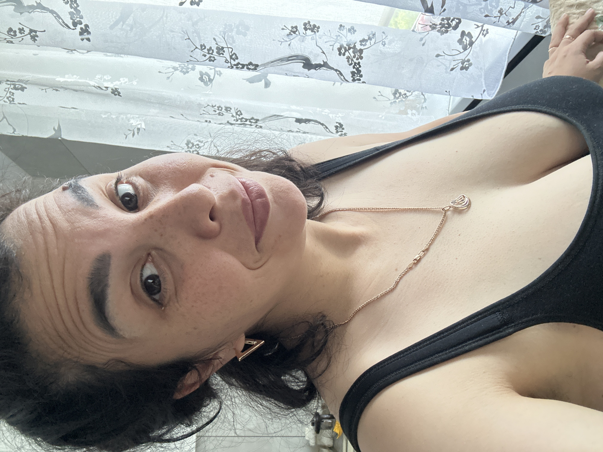 The image shows a close-up of a woman from the chest up, posing for a selfie in front of a window with decorative curtains. The woman has dark hair pulled back and is wearing a black tank top, a rose gold necklace with a pendant, and triangular earrings. She has a light skin tone with freckles and a mole above her left eyebrow. The background consists of a white sheer curtain with a floral pattern of trees and birds, and a hint of natural light coming through the window. The environment appears to be indoors, possibly a home.