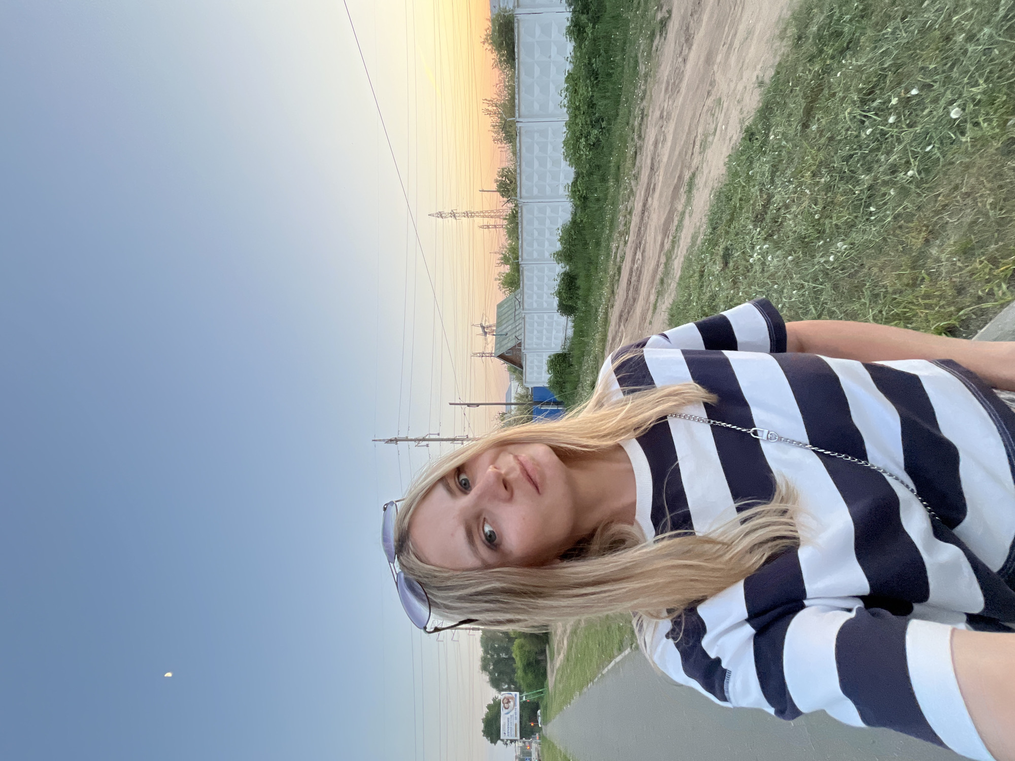 A young woman poses for a photograph on a road in Chaikovsky, Russia. She is wearing a striped navy blue and white shirt and has blonde hair. Sunglasses are perched on her head. In the background, there are electrical power lines, a fence, a dirt path, grass, and a building with a blue tarp. The sky transitions from blue to orange in the evening light. A pale moon is visible in the sky. An advertisement sign is visible in the distance.