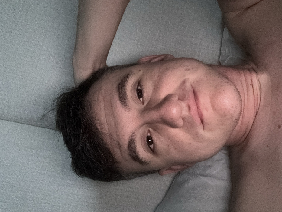 An adult male is depicted in a close-up, eye-level indoor shot. He has short dark hair, fair skin, and is looking directly at the camera with a neutral expression transitioning into a subtle smile. His right arm, from the viewer's perspective, is bent with the hand positioned behind his head. He appears shirtless, with a bare left shoulder visible in the lower left quadrant of the frame. He is positioned against a light blue-grey, textured fabric surface, likely a pillow or upholstered furniture. The ambient lighting is dim, creating soft shadows, particularly noticeable under the chin and along the right side of his face. The visual information available in the image does not permit the identification of a specific location within the city of Kastelruth - Castelrotto, Italy.