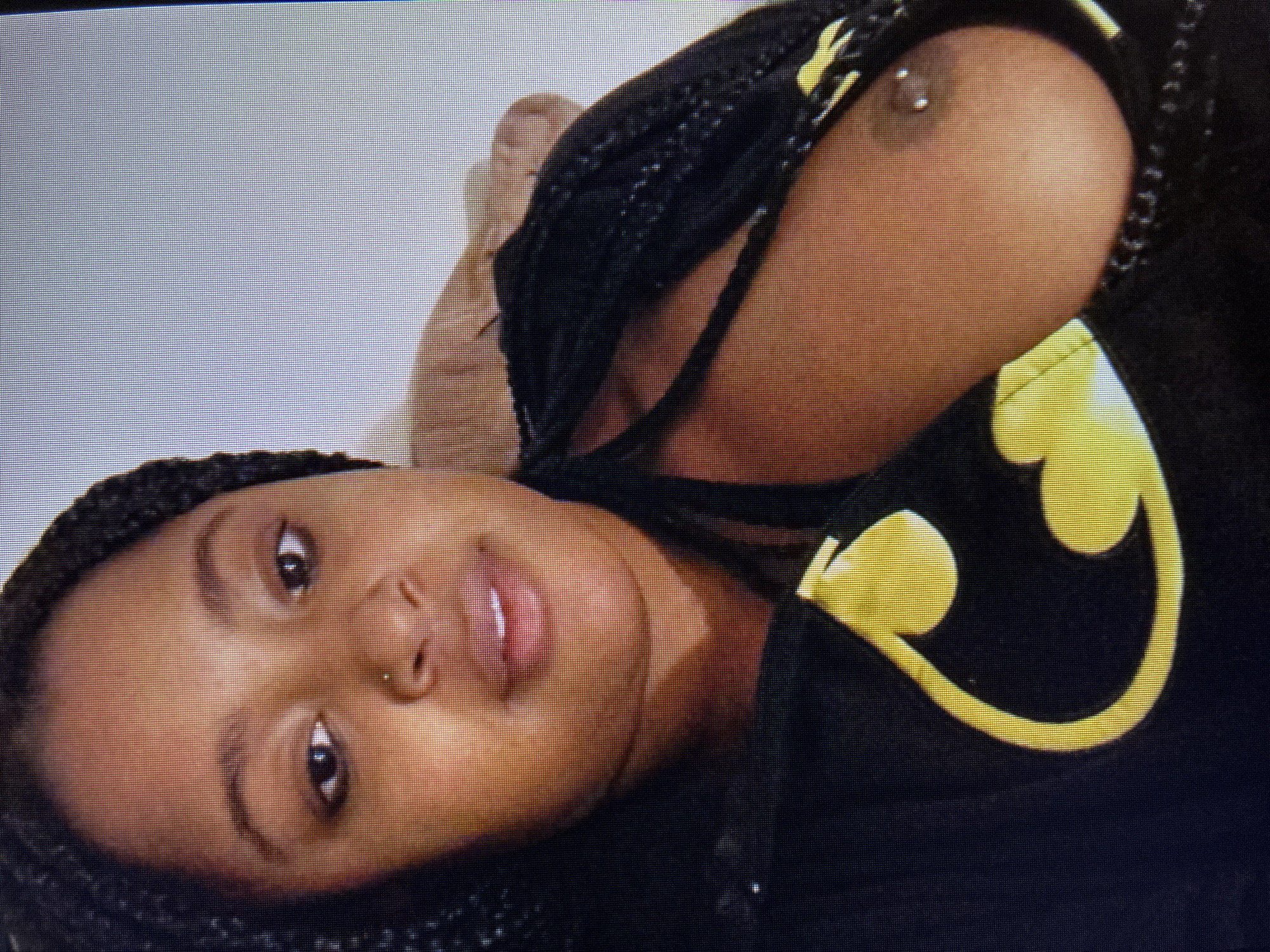 The media file presents a close-up, portrait-style image of a person, likely female, with dark skin and braided hair, captured in a reclining position. The individual is looking directly towards the camera with a neutral expression, their head positioned towards the left of the frame.
They are wearing a dark, possibly black, top or hoodie, which features a prominent yellow logo resembling the Batman symbol on the chest area. A piercing is visible near their nose. Their left breast is partially exposed, revealing a nipple piercing. The lips are slightly parted. The background is a plain, light, and unadorned surface, indicating an indoor setting. There are no specific visual cues to determine the time of day, weather, or any particular location within Varna, Bulgaria. The image itself appears to be a photograph of a digital screen, as indicated by the visible pixel grid throughout.